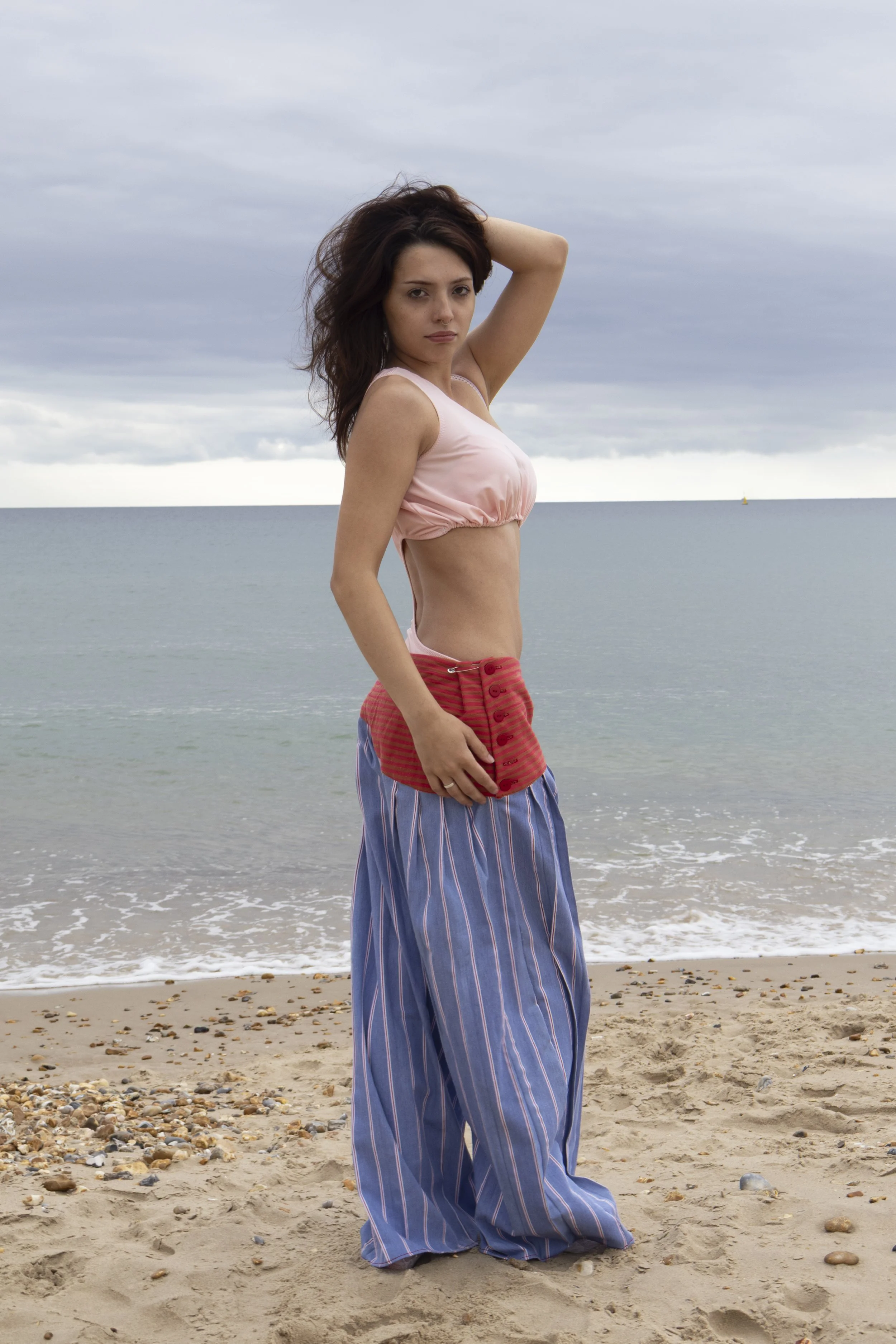 Woman standing on a sandy beach with small rocks, ocean and cloudy sky in the background. She has long, wavy brown hair, is wearing a pink crop top and striped wide-leg pants.