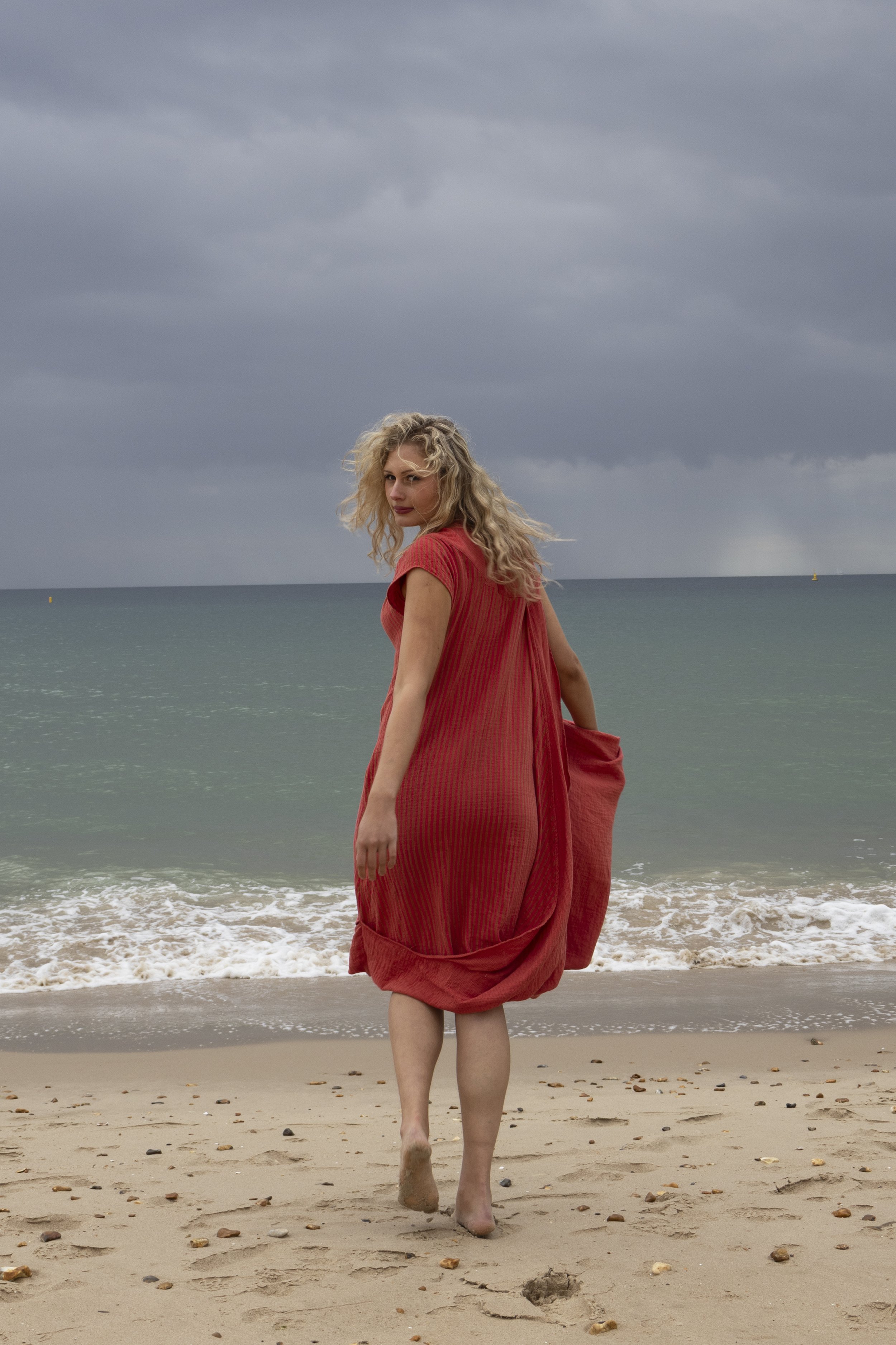 A woman in a red dress walking barefoot on a sandy beach with ocean waves and a cloudy sky in the background.