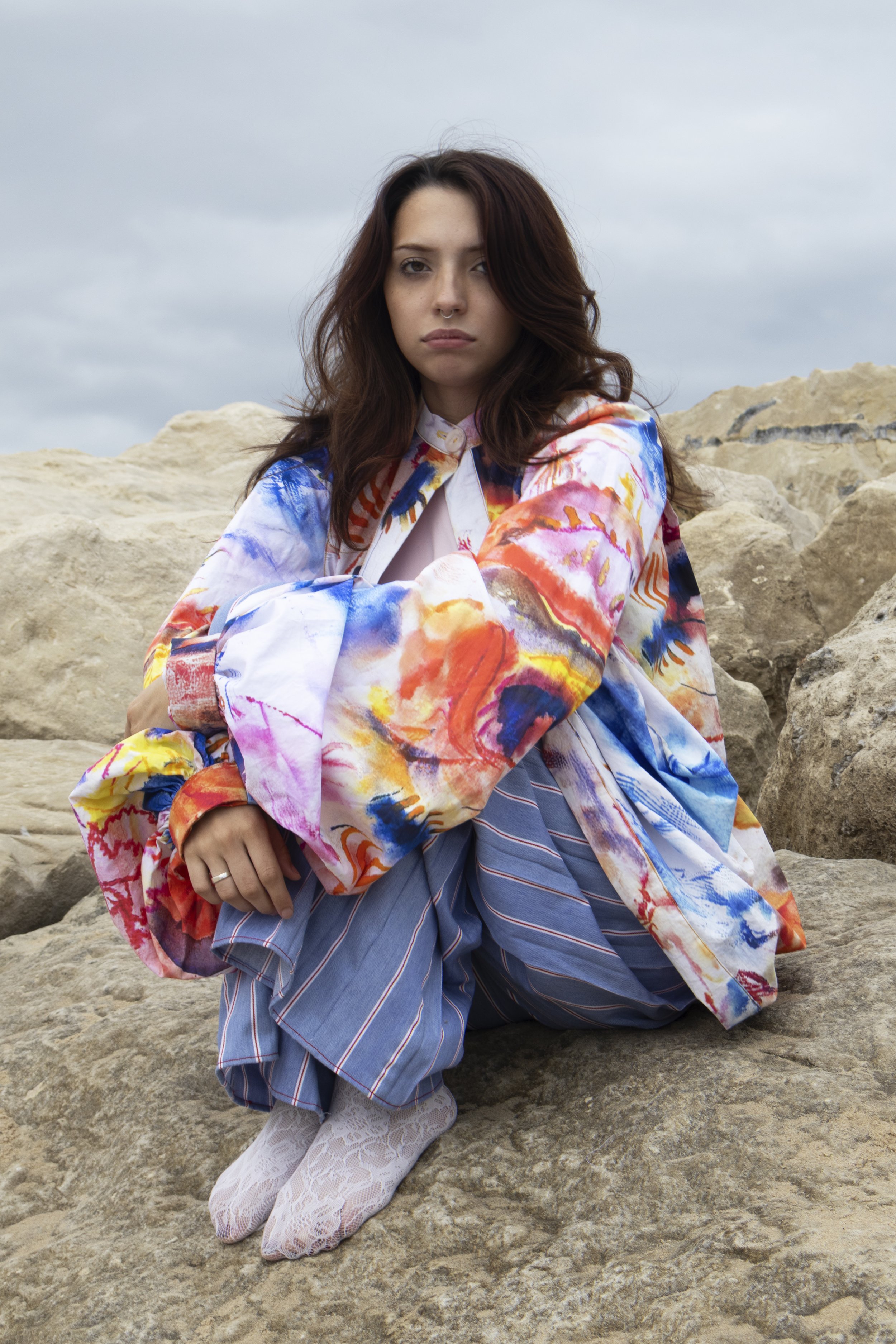 Young woman sitting on rocks outdoors, wearing a colorful, abstract-patterned jacket, striped pants, and lace tights, with a cloudy sky in the background.