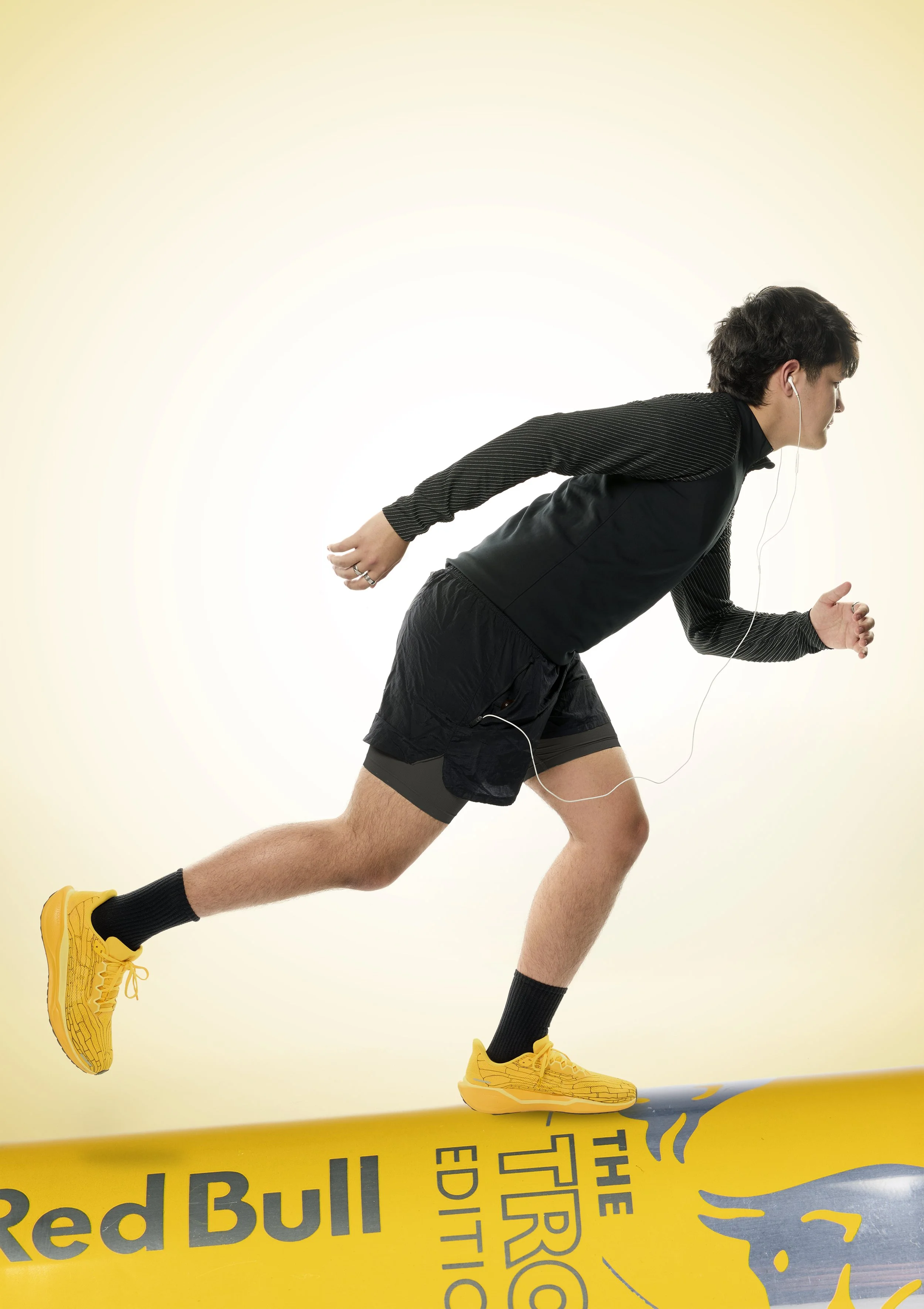 A man running on a yellow Red Bull energy drink platform wearing yellow running shoes, black shorts, a black long sleeve shirt, and earphones.