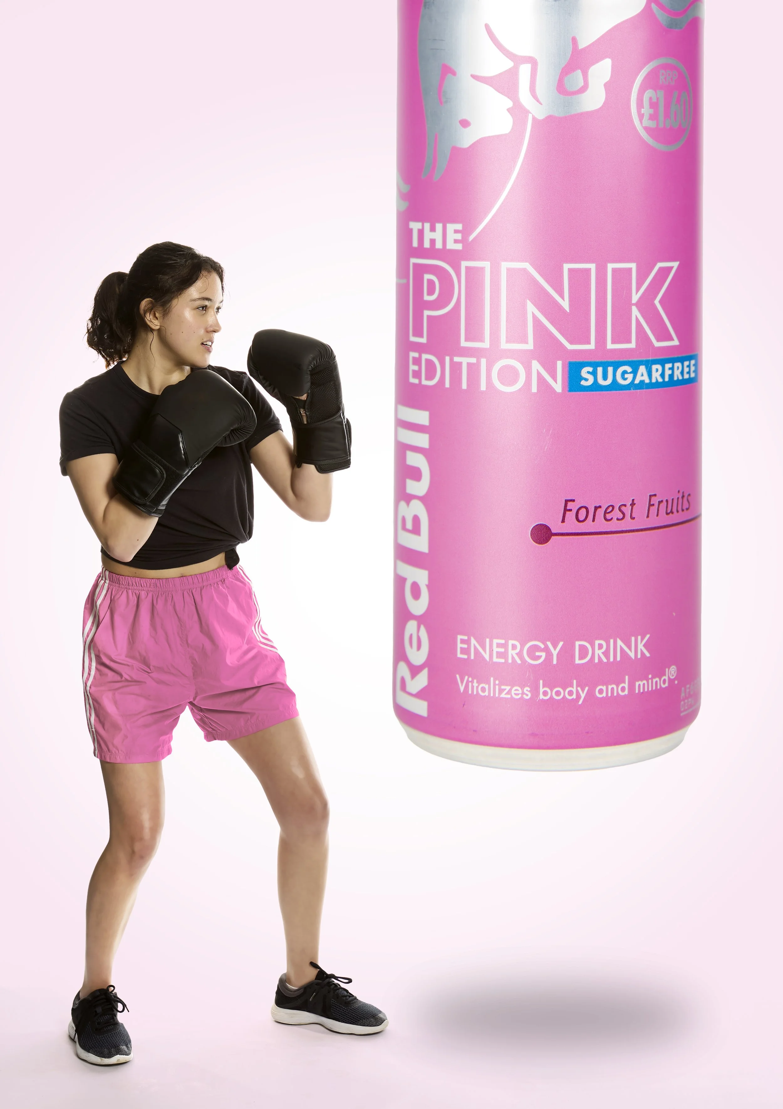 A woman in athletic clothing wearing boxing gloves, standing in a fighting pose next to a large pink energy drink can labeled 'The Pink Edition Sugarfree Forest Fruits' on a light pink background.