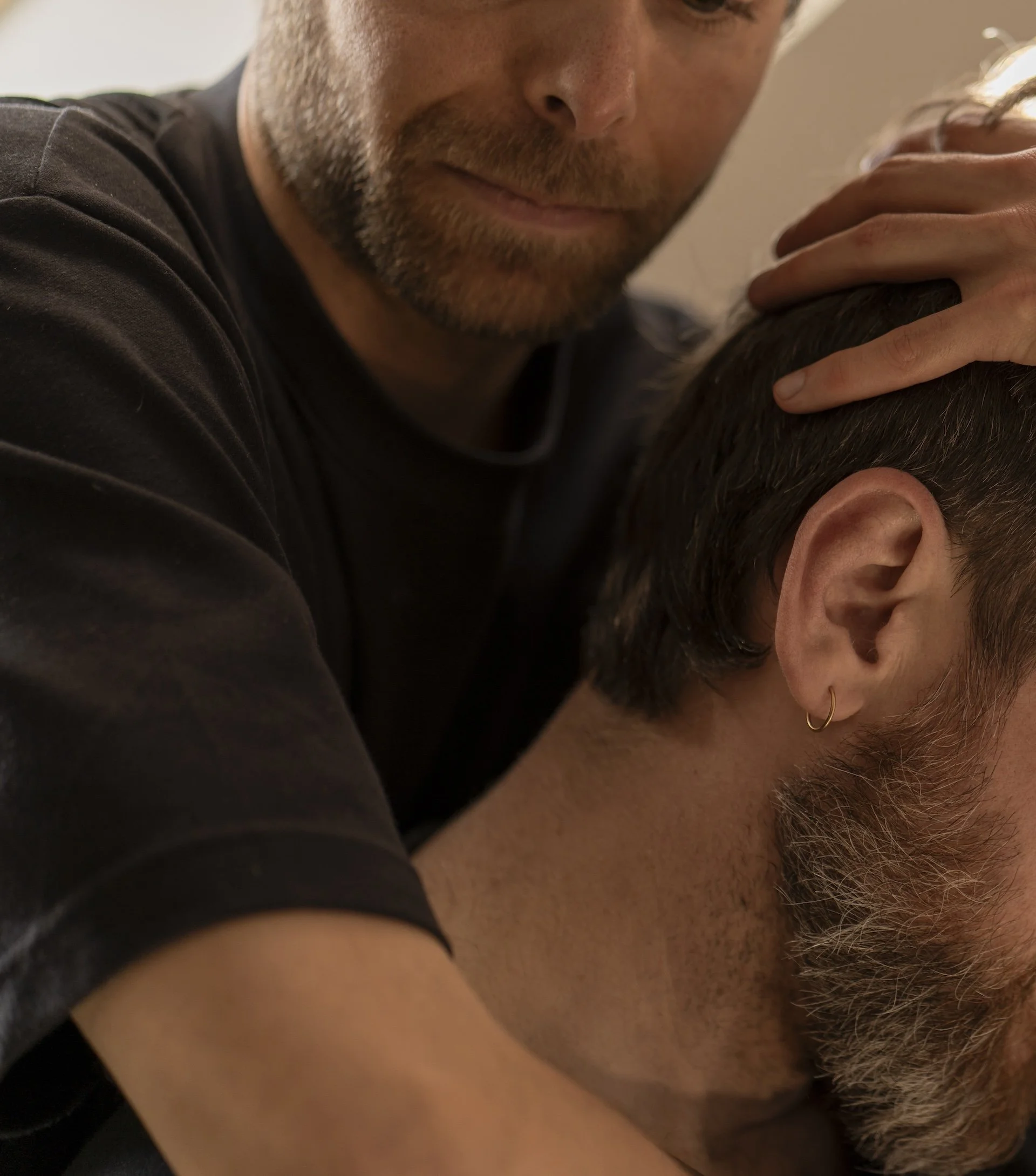 Un homme avec une barbe est allongé avec sa tête reposant contre une personne à cheveux foncés et courts, dont la tête est inclinée vers le bas. L'homme regarde vers le haut avec une expression douce.