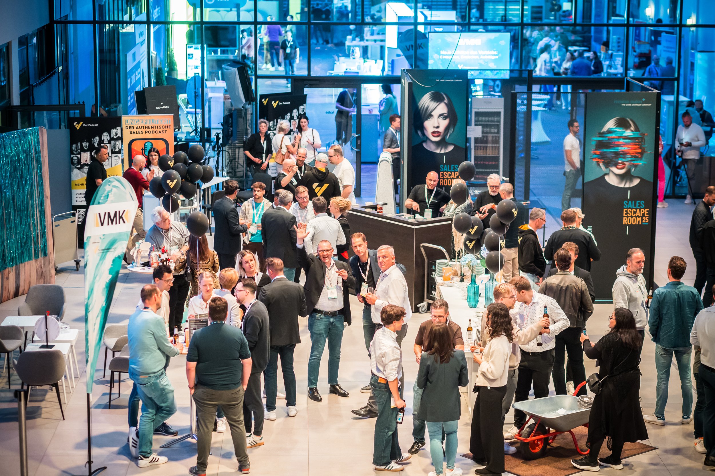 Bilder von einer Messehalle mit vielen Menschen, die sich an verschiedenen Ständen versammeln, darunter einen Getränke- und einen Informationsstand, mit großen Plakaten und schwarzen Ballons, im Hintergrund große Bildschirme und glasartige Wände.