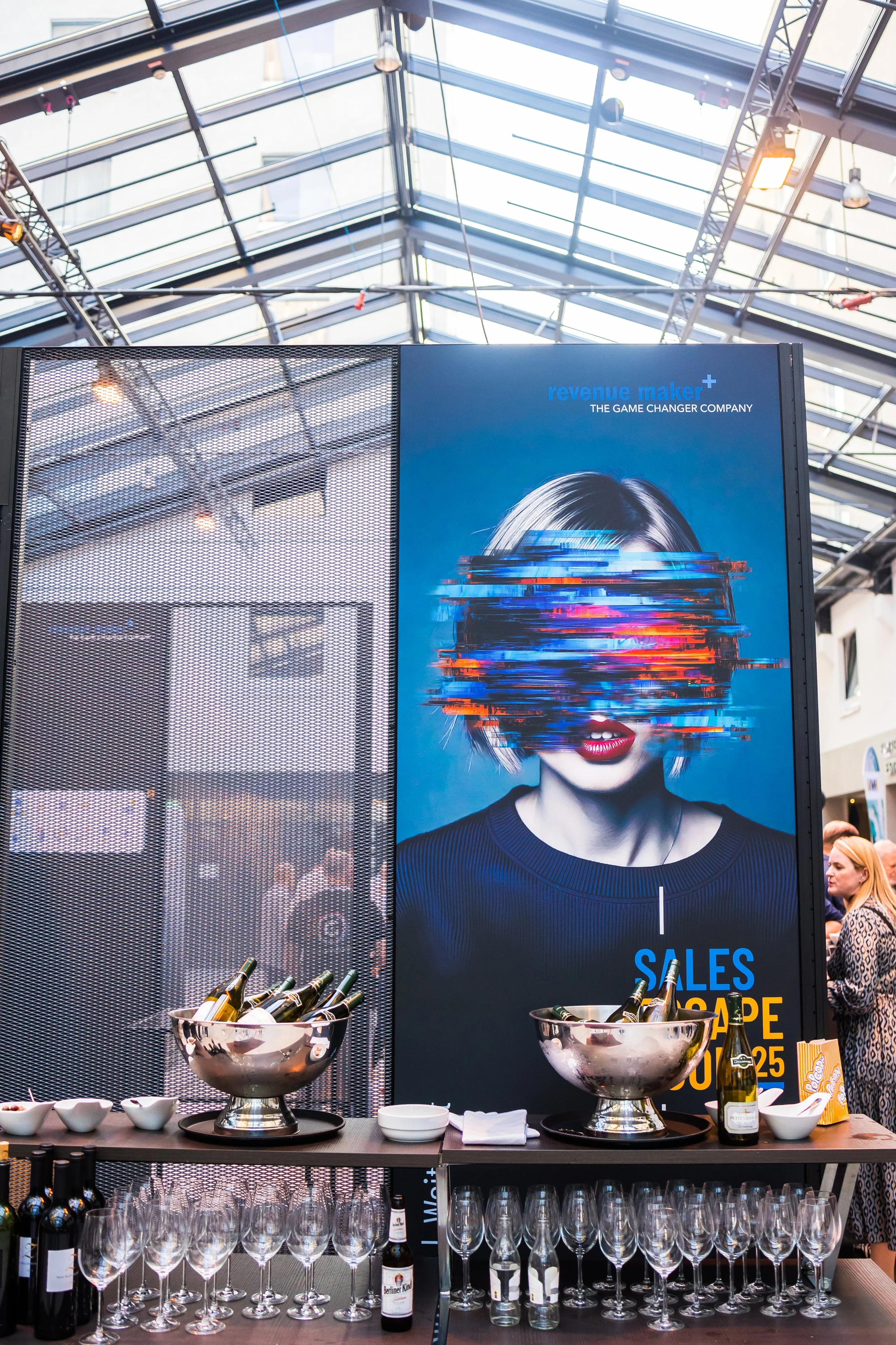 Ein Ausstellungsstand mit einem großen Plakat, das eine Frau mit gestörtem Gesicht zeigt. Vor dem Stand stehen Gläser, Weinflaschen und eine große Chips-Tüte, vermutlich bei einer Verkaufsveranstaltung oder Messe.