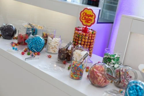 Verschiedene Süßigkeiten in Behältern auf einem weißen Tisch, darunter Gummibärchen und Bonbons, mit einem Chupa Chups Logo.