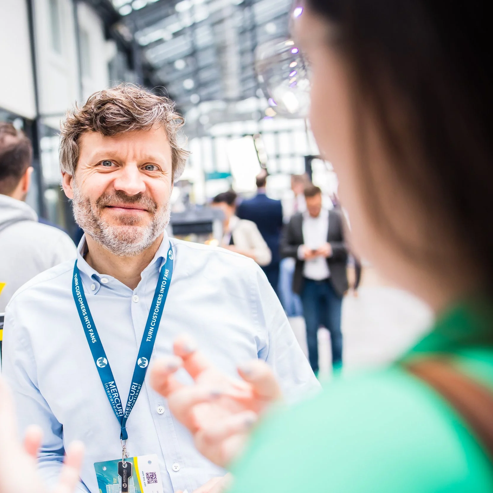 Mann mit grauem Bart im Gespräch auf einer Messe, Getränk in der Hand, mit anderen Menschen im Hintergrund.