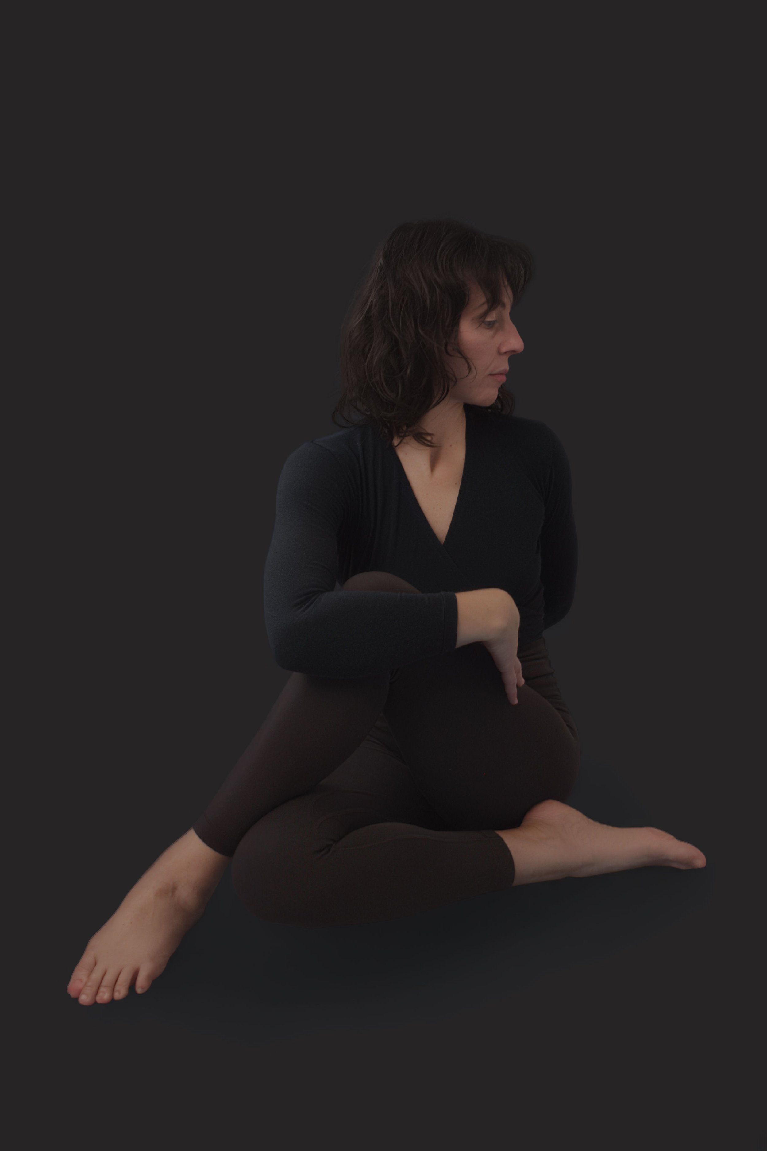 A woman with dark, wavy hair in a yoga pose against a black background, wearing a black long-sleeve top and black leggings, with Earth tones in her makeup.