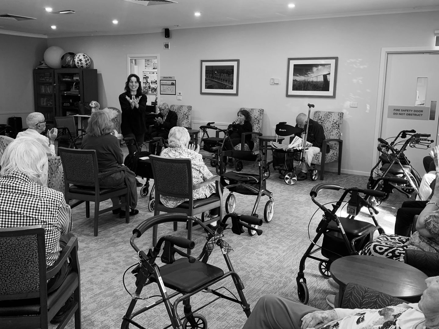 A group of elderly people, some with walkers, attending a performance or activity in a common room. A woman is standing at the front, talking or performing, while others are seated and watching.