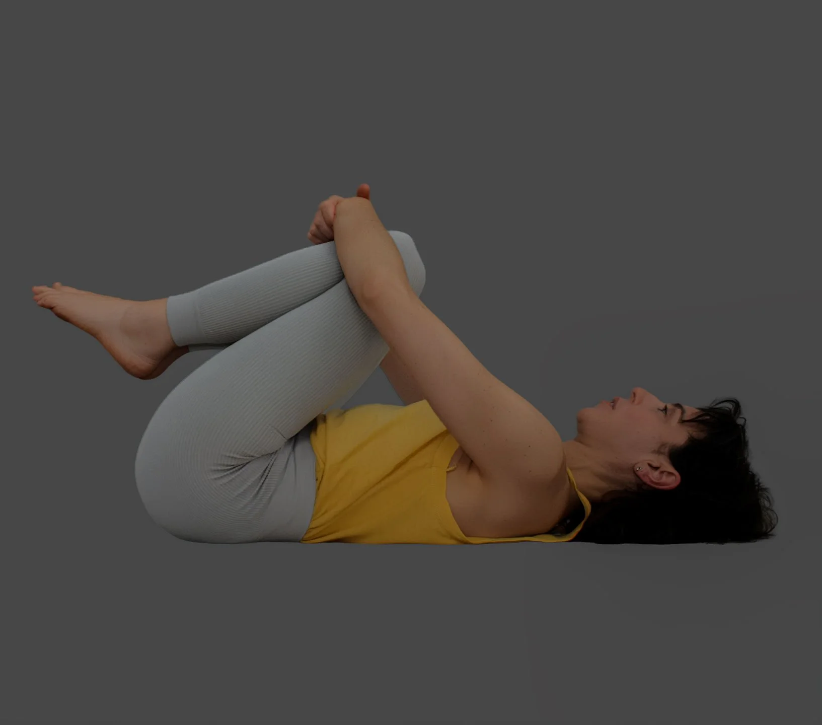 A woman practicing a yoga pose called Happy Baby, lying on her back with her knees bent towards her chest, holding her feet with her hands.