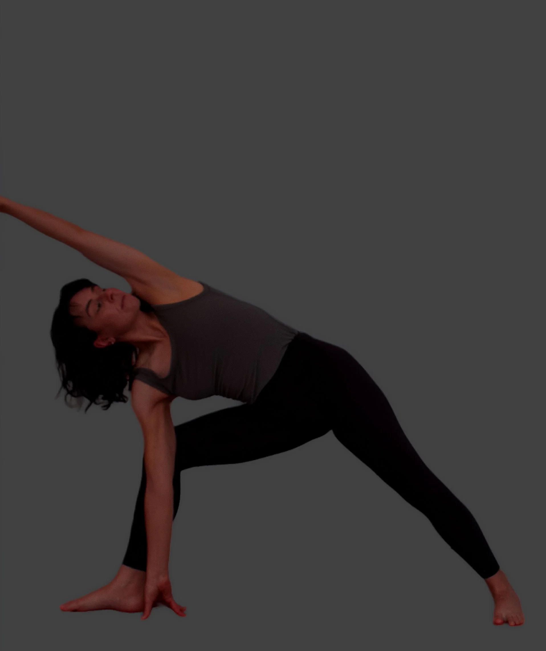 A woman performing a yoga pose called triangle pose, standing with legs apart, one arm stretching upward, and the other arm reaching toward her foot, against a gray background.
