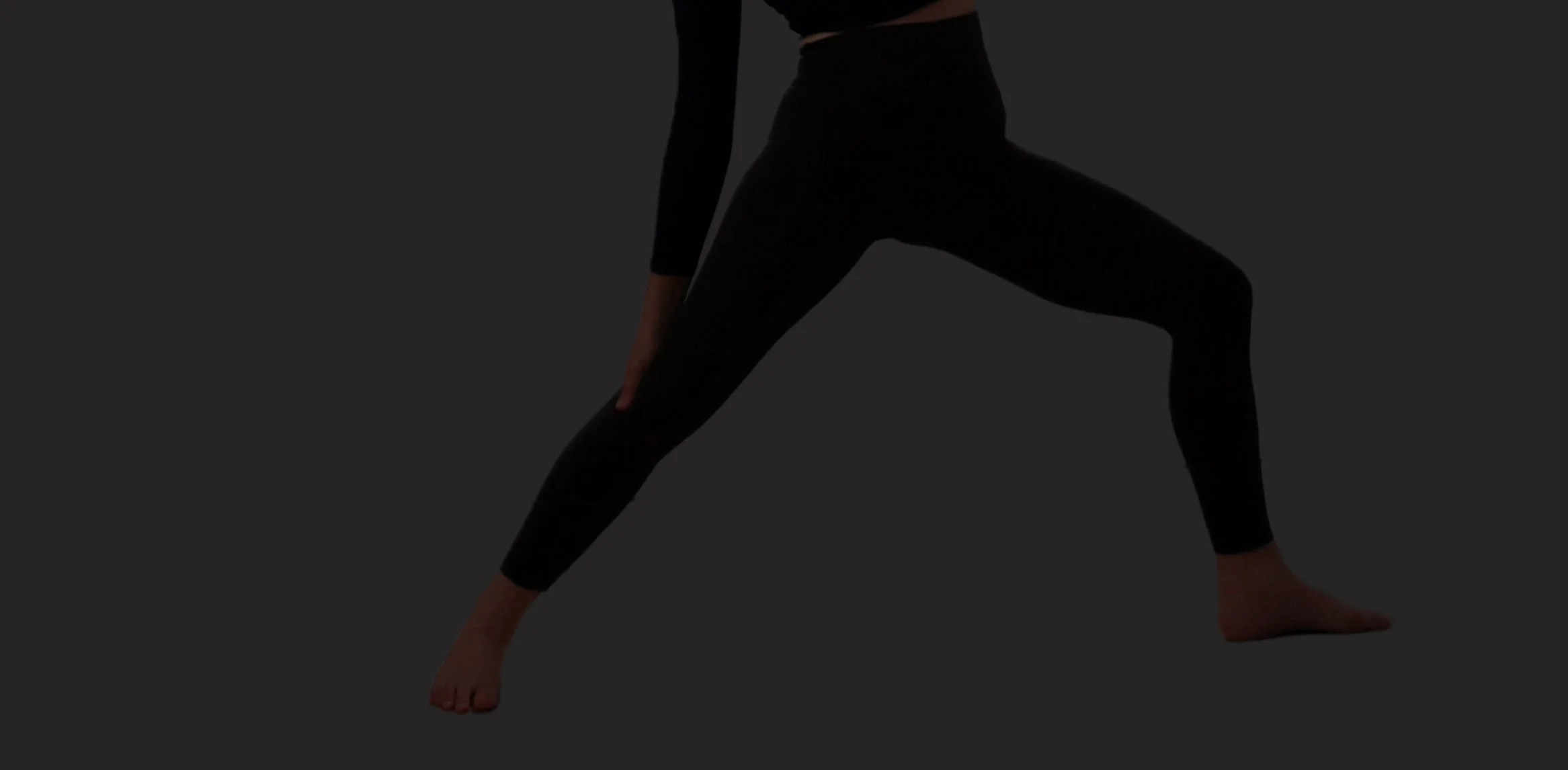 A person in black clothing performing a yoga pose against a dark background, with one hand on the floor and the other arm bent and resting on the bent knee.