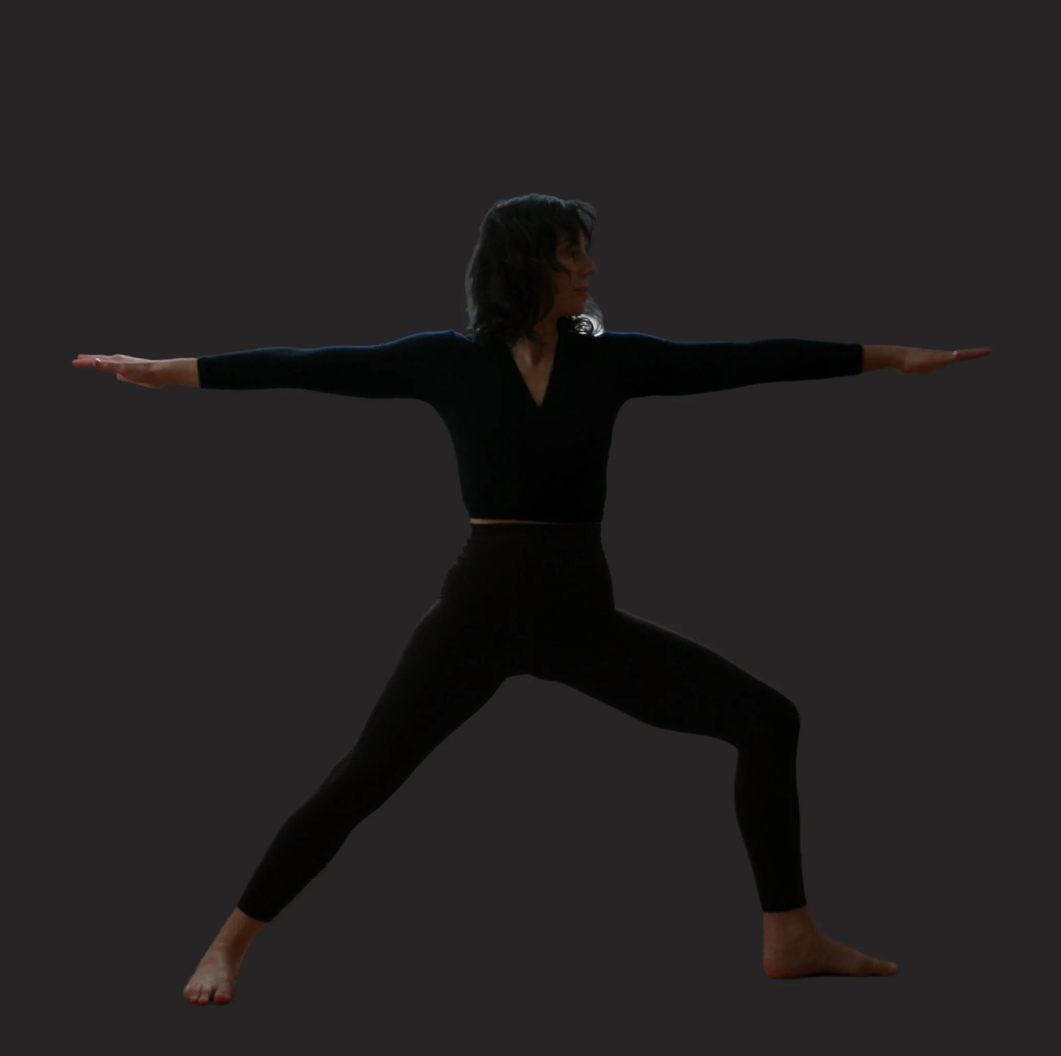 A woman in black athletic clothing performing a yoga pose with arms extended horizontally and legs in a lunge position against a dark background.