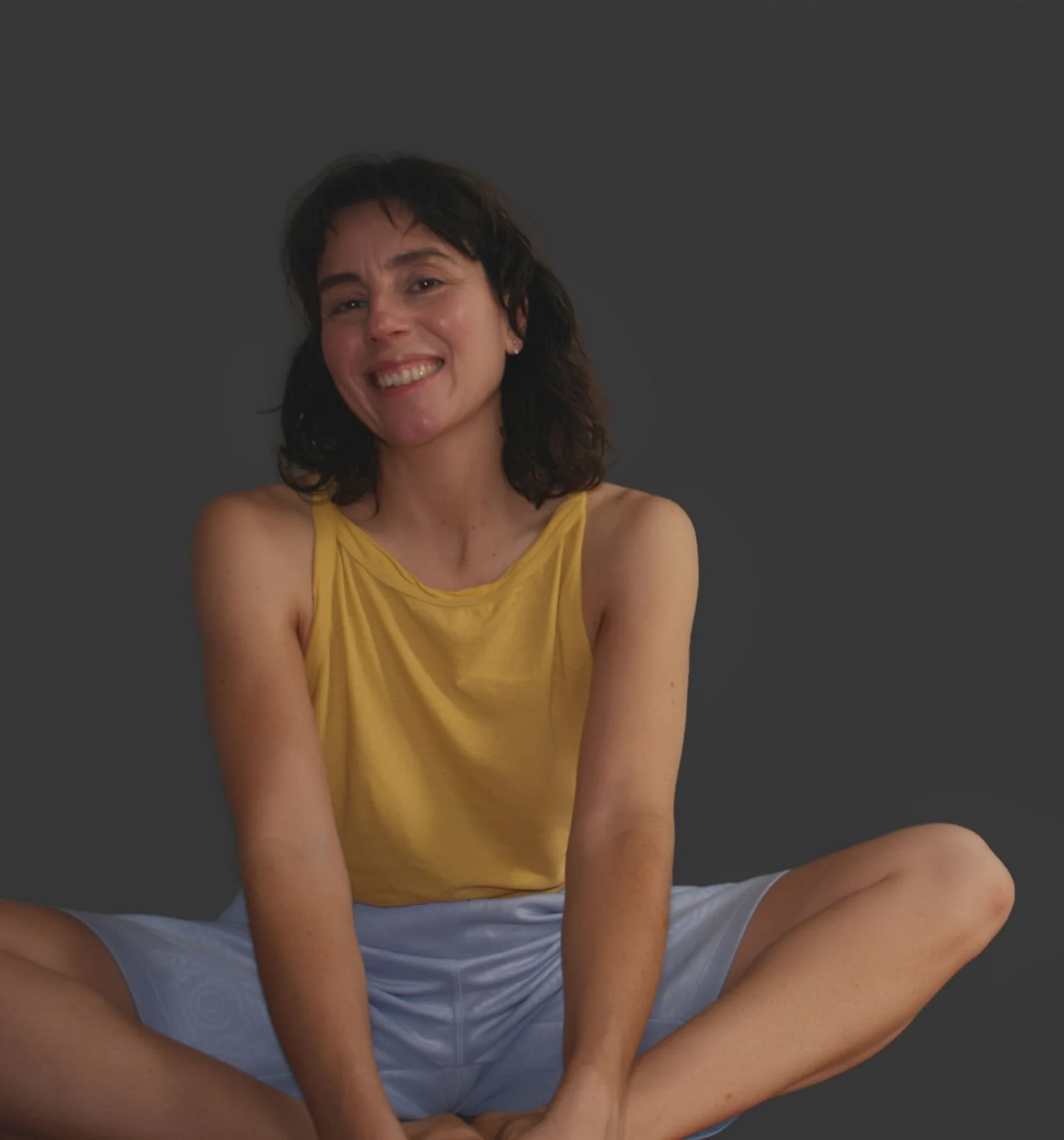 A woman smiling, sitting cross-legged against a dark gray background, wearing a yellow sleeveless top and light-colored shorts.