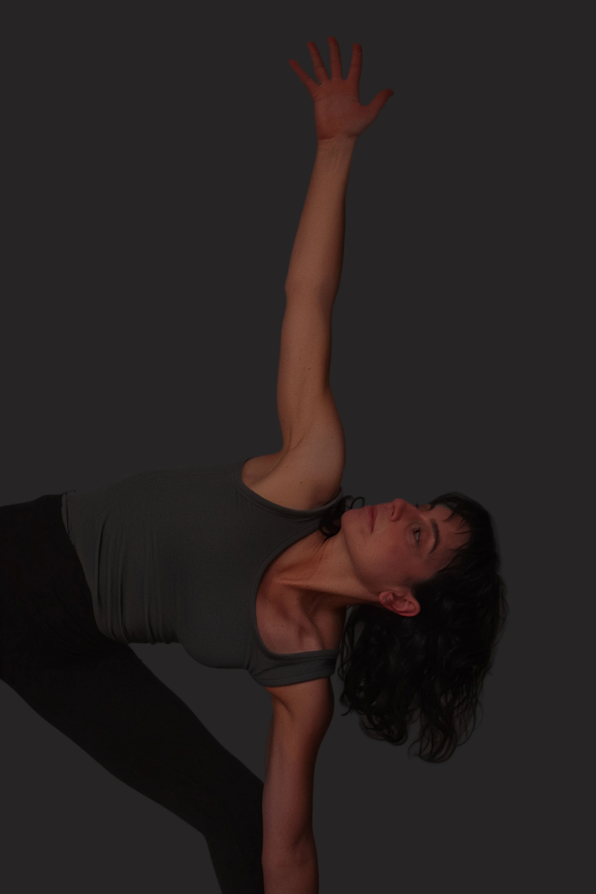 A woman practicing yoga in a pose, lying on her side with one arm extended upward and the other supporting her body, wearing a dark gray shirt and black pants.