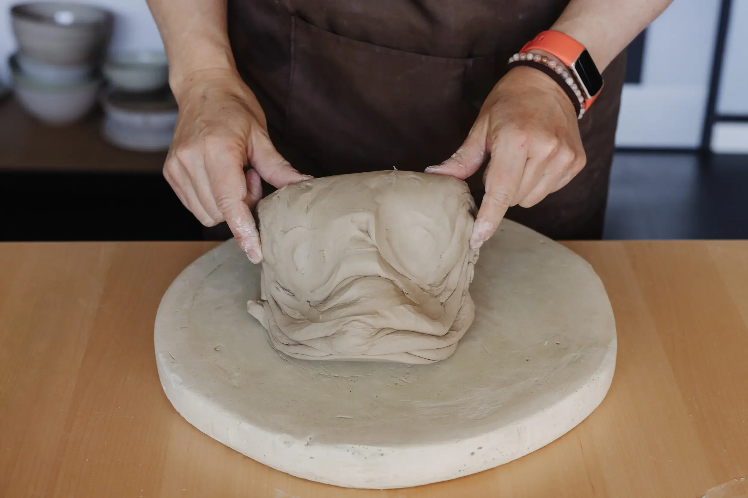 Handarbeit an einer Skulptur aus Ton auf einer runden Plattform. Hände modellieren die Skulptur, im Hintergrund sind Keramikschalen zu sehen.