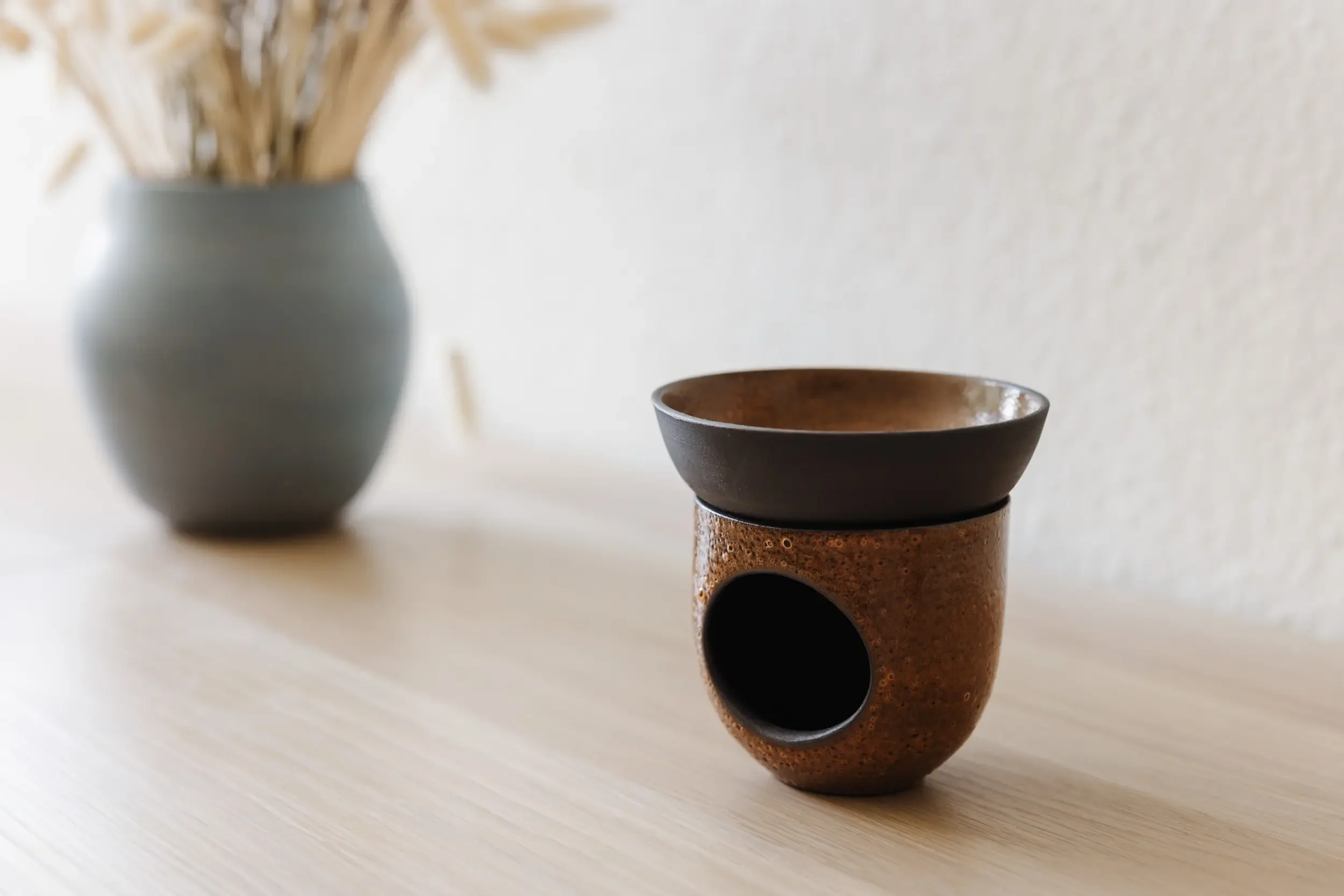 Zwei Keramikvasen, eine graue im Hintergrund mit trockenen Pflanzen, und eine braune mit schwarzer Öffnung vorne auf einem Holztablett