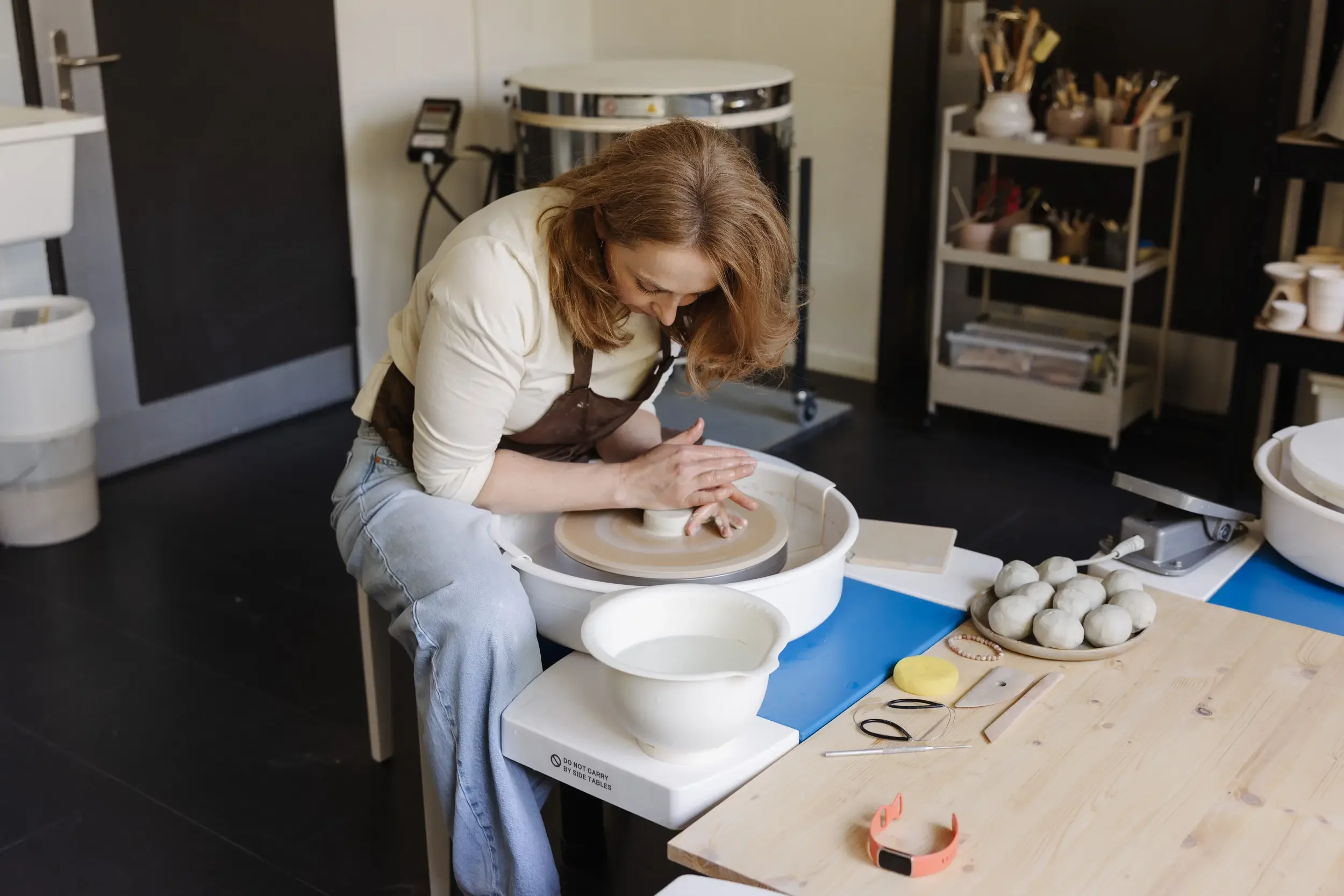 Eine Frau formt Ton auf einer zweistufigen Töpferscheibe in einer Werkstatt. Im Hintergrund Regale mit Töpferwerkzeugen und Keramiken, auf dem Tisch befinden sich verschiedene Werkzeuge und fertige Keramikstücke.