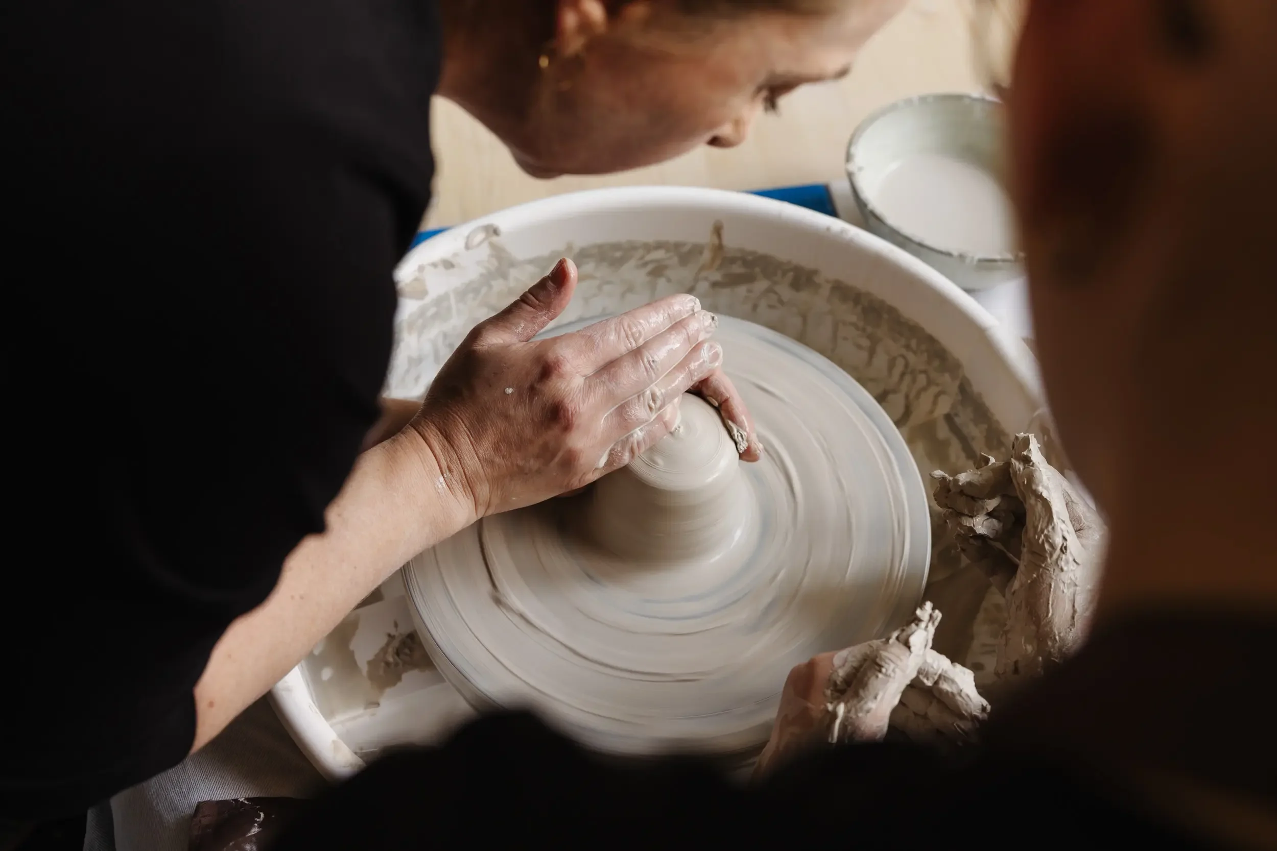Eine Frau arbeitet an einer Töpferscheibe und formt eine Vase aus Ton. Sie trägt eine schwarze Kleidung und hat kurze, blond gefärbte Haare. Der Arbeitsbereich ist mit Ton und Skulpturen bedeckt.