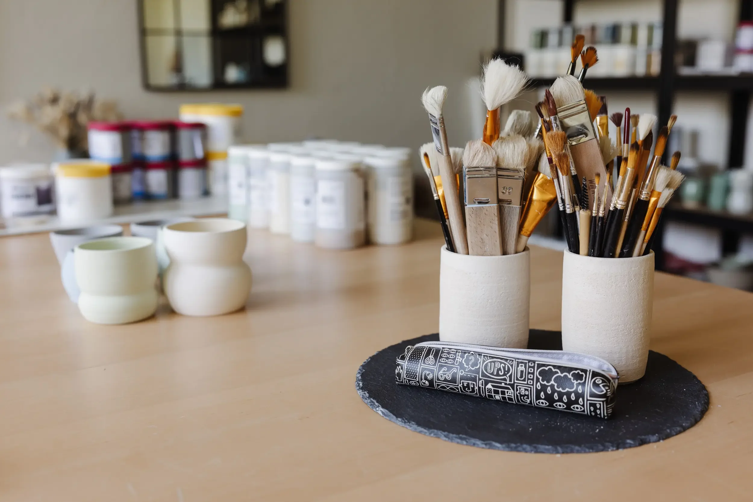 Pinsel in weißen Keramikvasen auf einem Tisch, im Hintergrund verschiedene Farben und Kunstmaterialien.