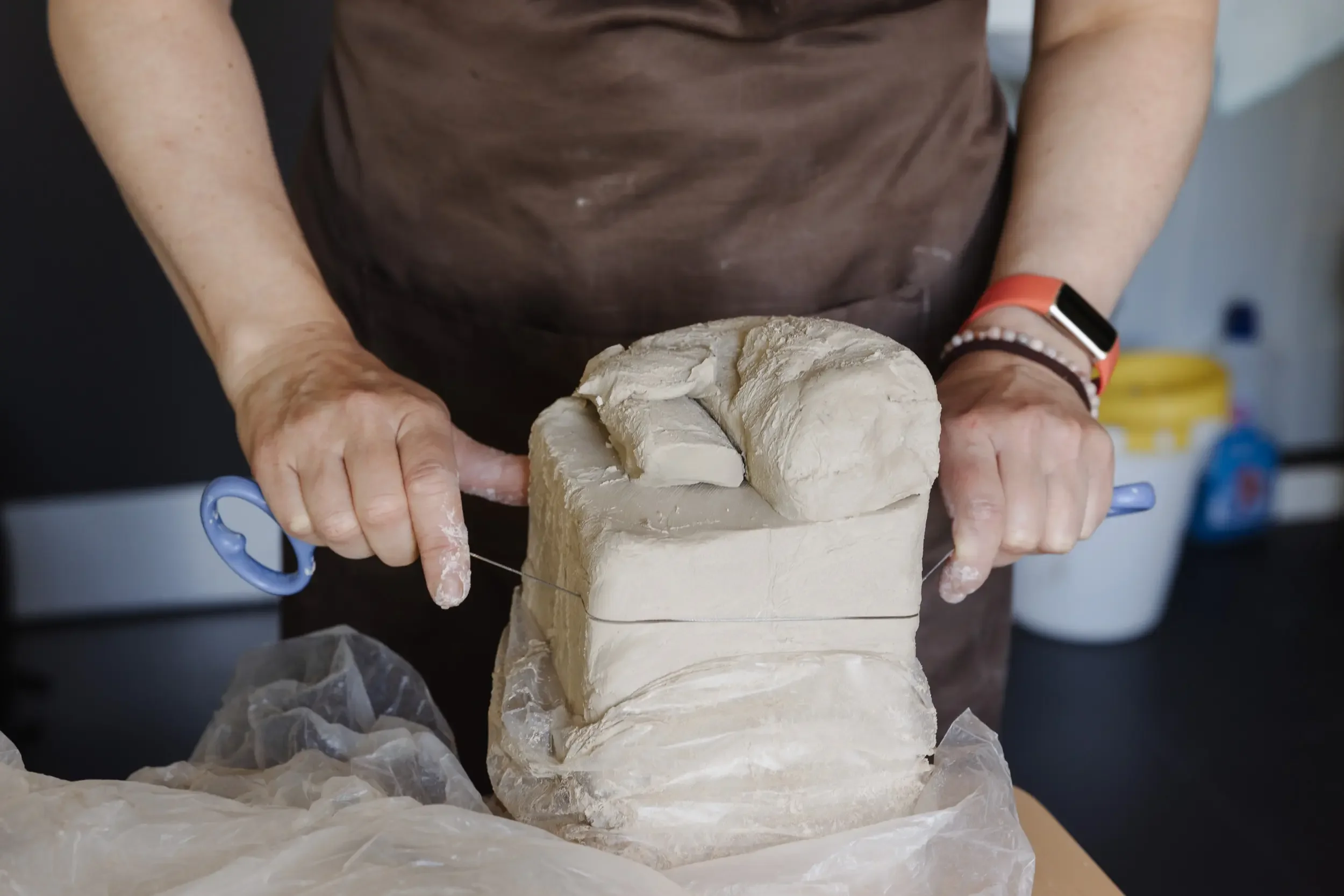 Person modelliert Skulptur aus Ton, mit Werkzeug in Hand, auf Tisch mit Abdeckpapier, Hintergrund mit blauen und gelben Gegenständen.