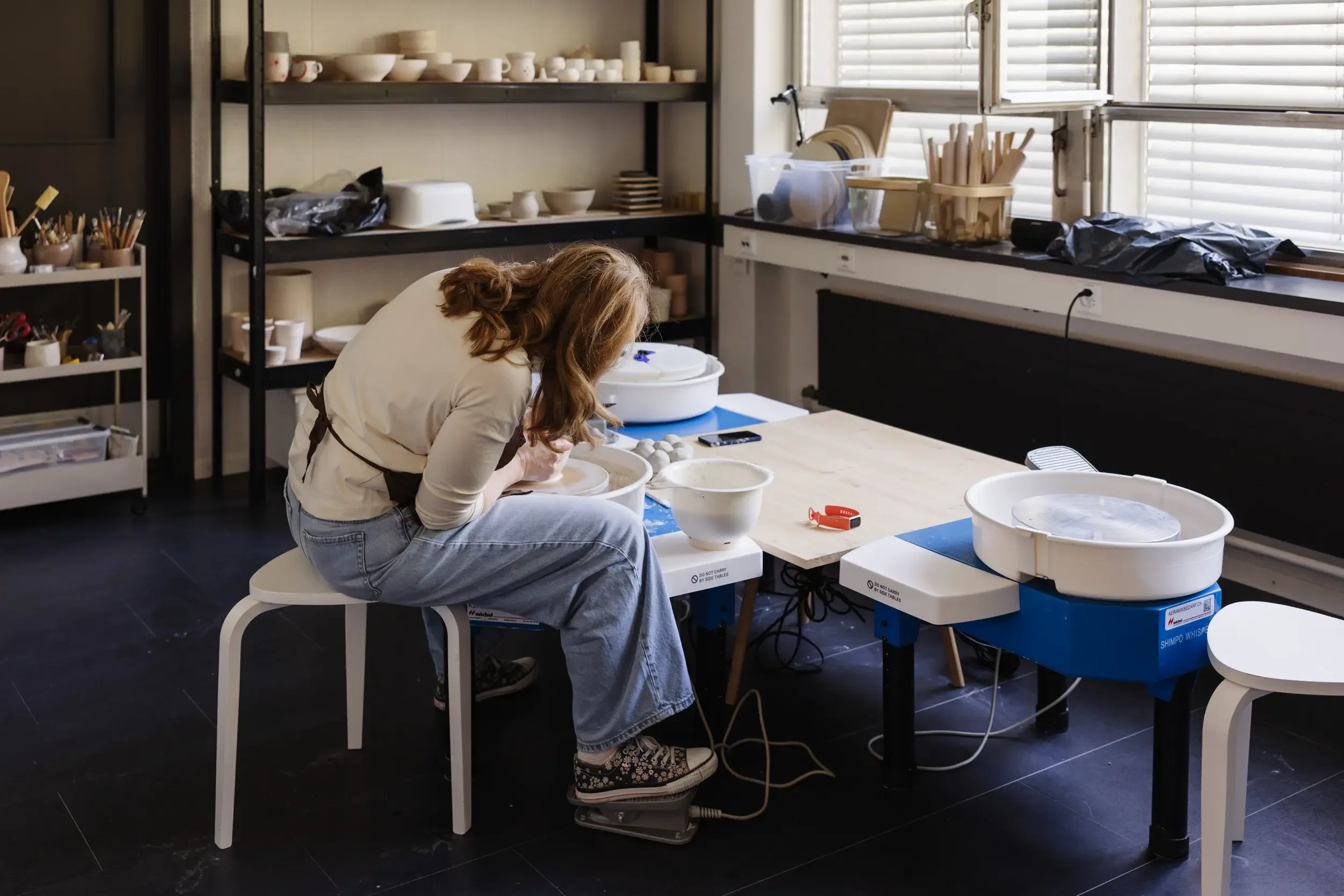 Eine Person arbeitet in einem Keramielabor, formt eine Tonware auf einer Töpferscheibe, umgeben von keramischem Werkzeug und Keramikstücken auf Regalen.