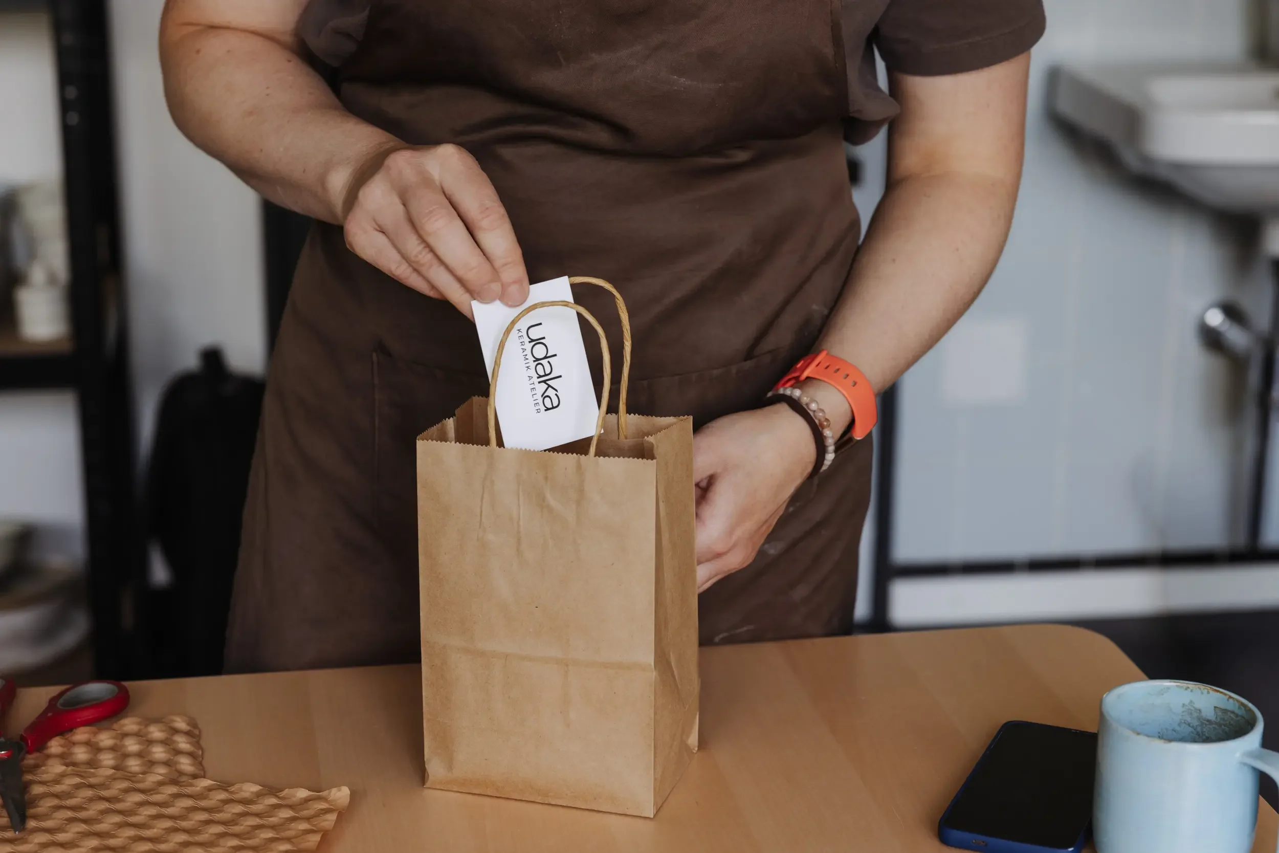 Eine Person hält einen kleinen Papiersack mit einem Etikett, das „UdaKa“ liest, wahrscheinlich eine Marke oder ein Geschäft. Die Person trägt einen braunen Hemd und eine rote Smartwatch. Im Hintergrund sind Küchenutensilien sichtbar.