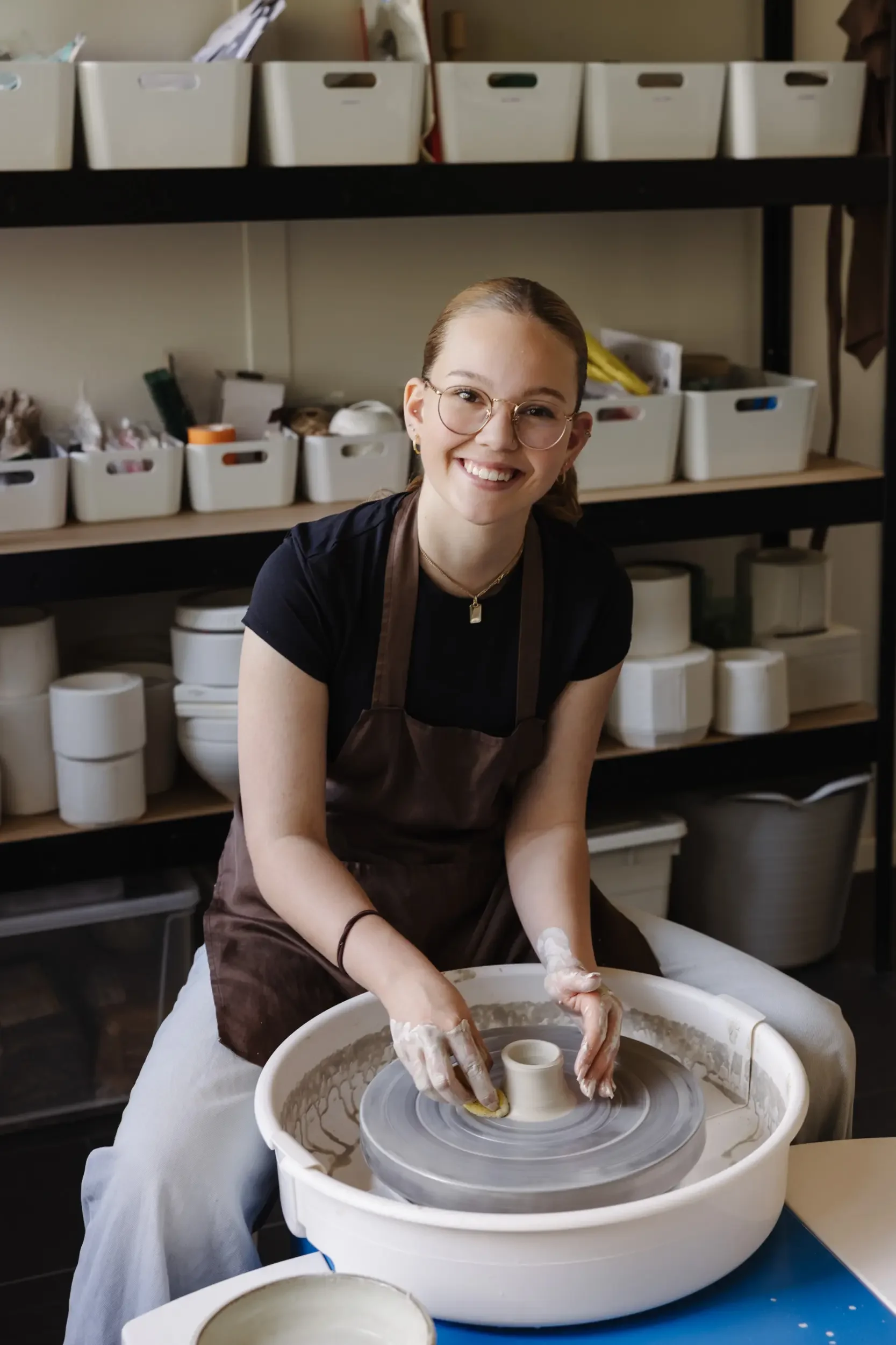 Junge Frau beim Töpfern in einer Keramikwerkstatt, sitzend an einer Töpfermaschine, lächelnd, trägt eine braune Schürze und Brille, im Hintergrund Regale mit Keramikstücken.