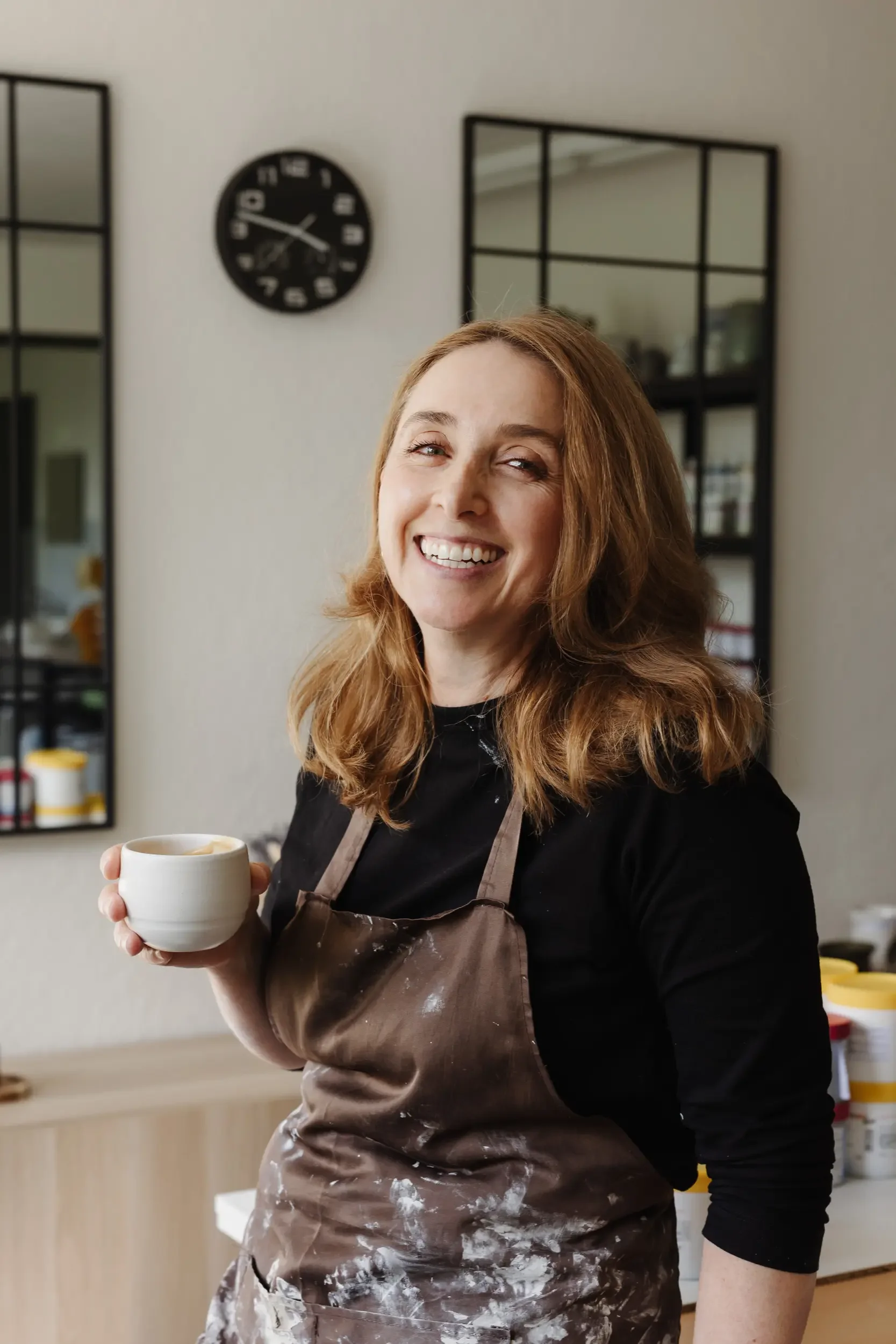 Besitzerin Kate in einer Schwarz- und Brauntracht hält eine weiße Tasse mit Kaffee, im Hintergrund sind Wände, Spiegel und Küchenutensilien sichtbar.