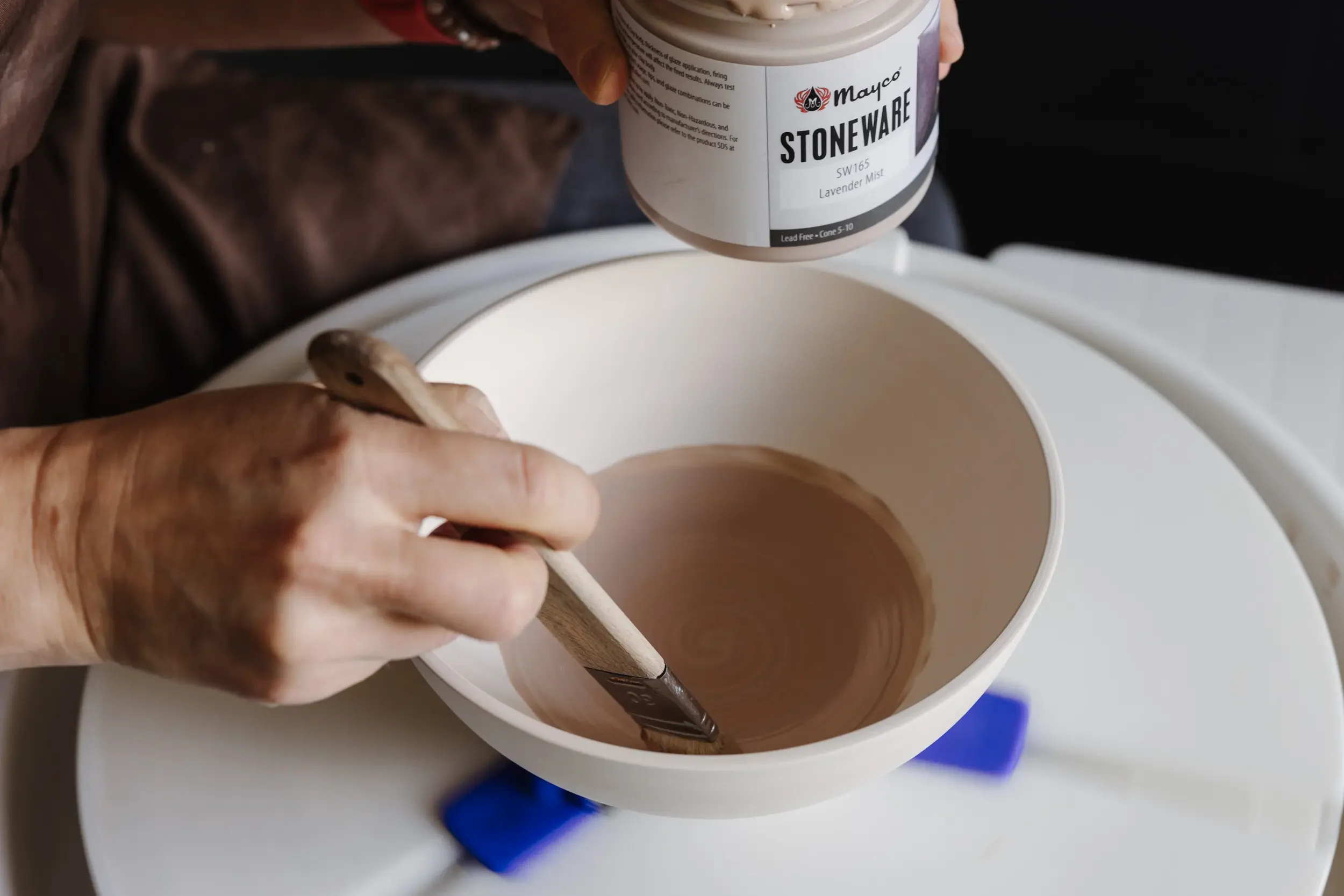 Person zeichnet mit einem Marker auf einem weißen Becher, während ein Farbkanister mit Lavendermist in der Hand gehalten wird.