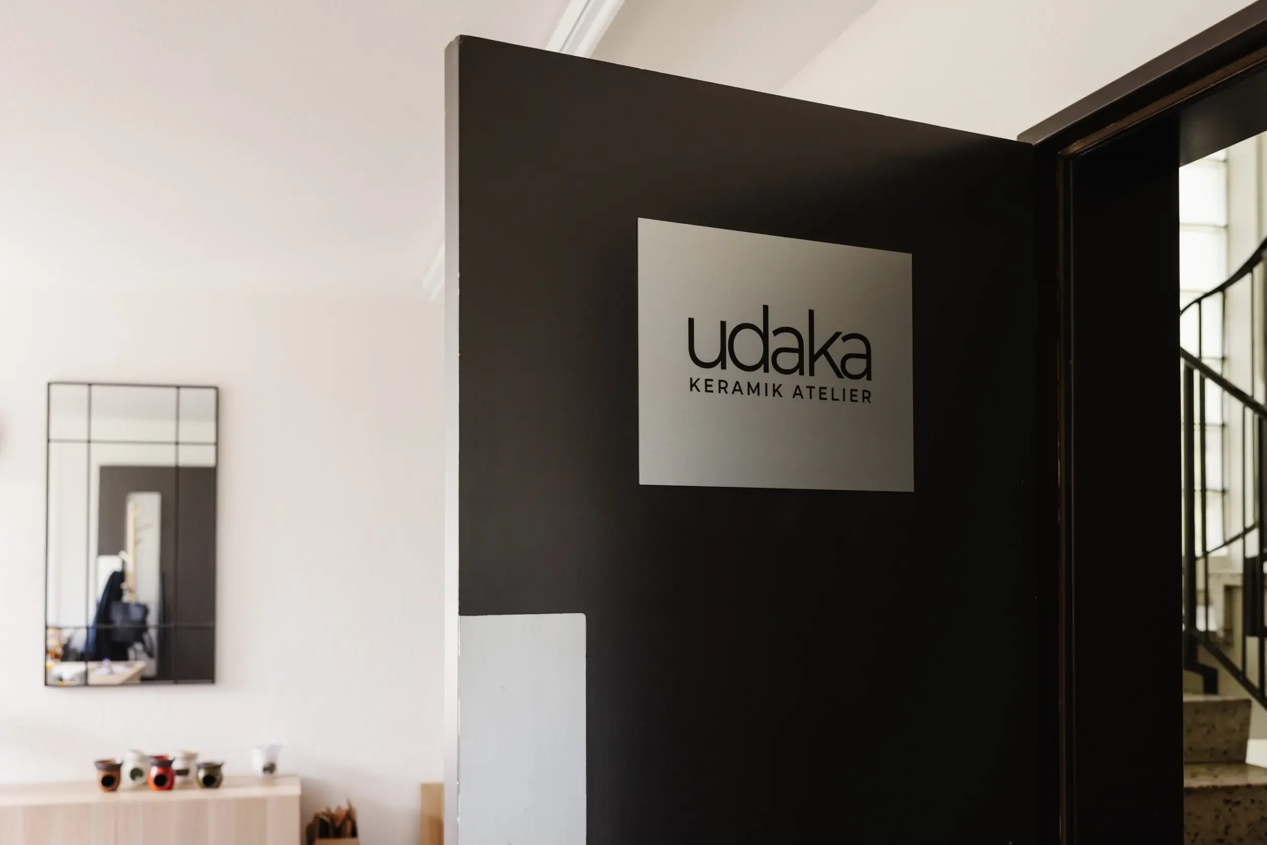 Innenraum eines keramischen Ateliers mit einer schwarzen Wand, auf der ein Schild mit der Aufschrift "udaka KERAMIK ATELIER" angebracht ist.