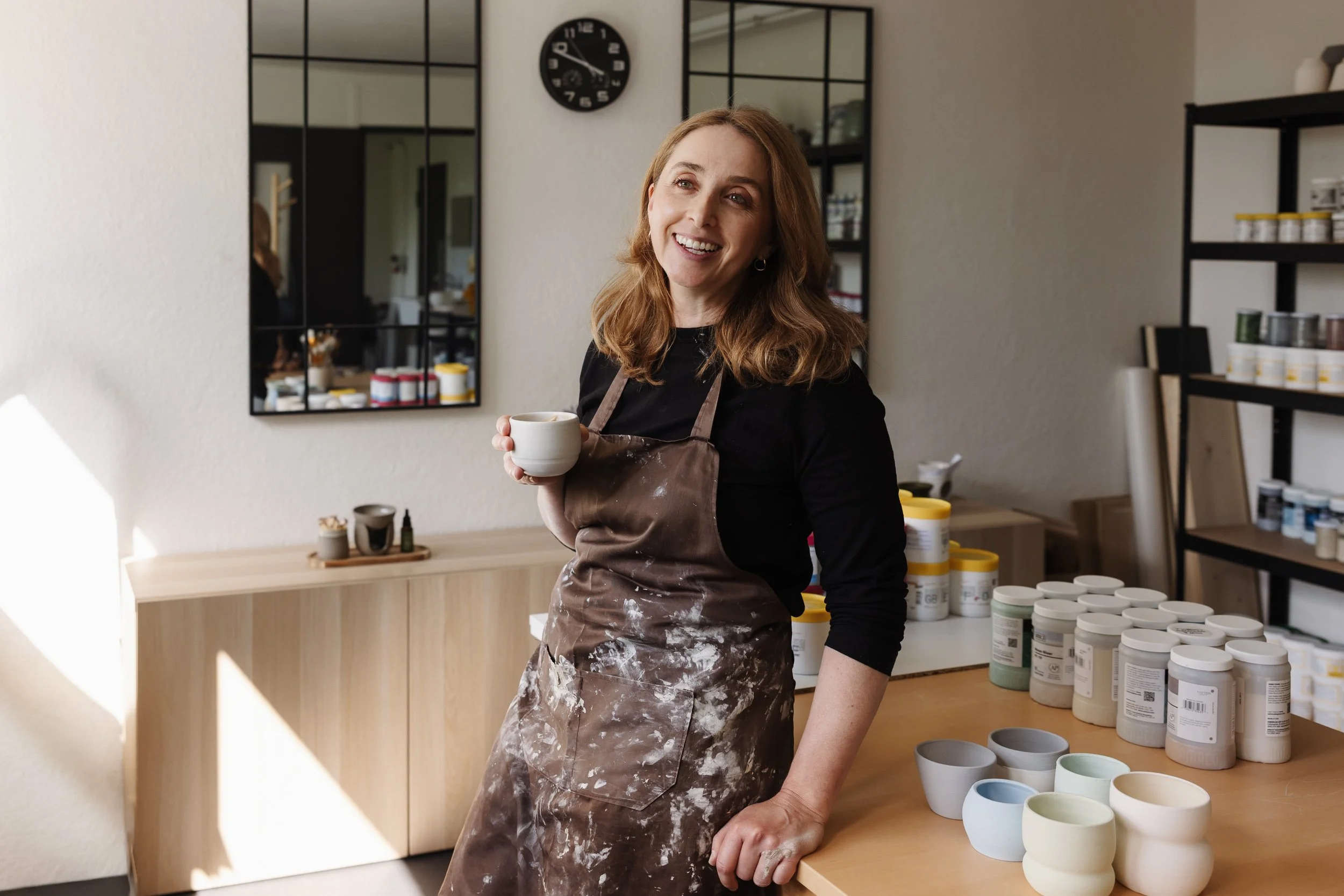 Eine Frau mit braunen Haaren und schwarzem Oberteil, die in einem Keramikstudio sitzt und eine weiße Tasse hält, umgeben von bunten Keramikgefäßen auf einem Tisch und Regalen.