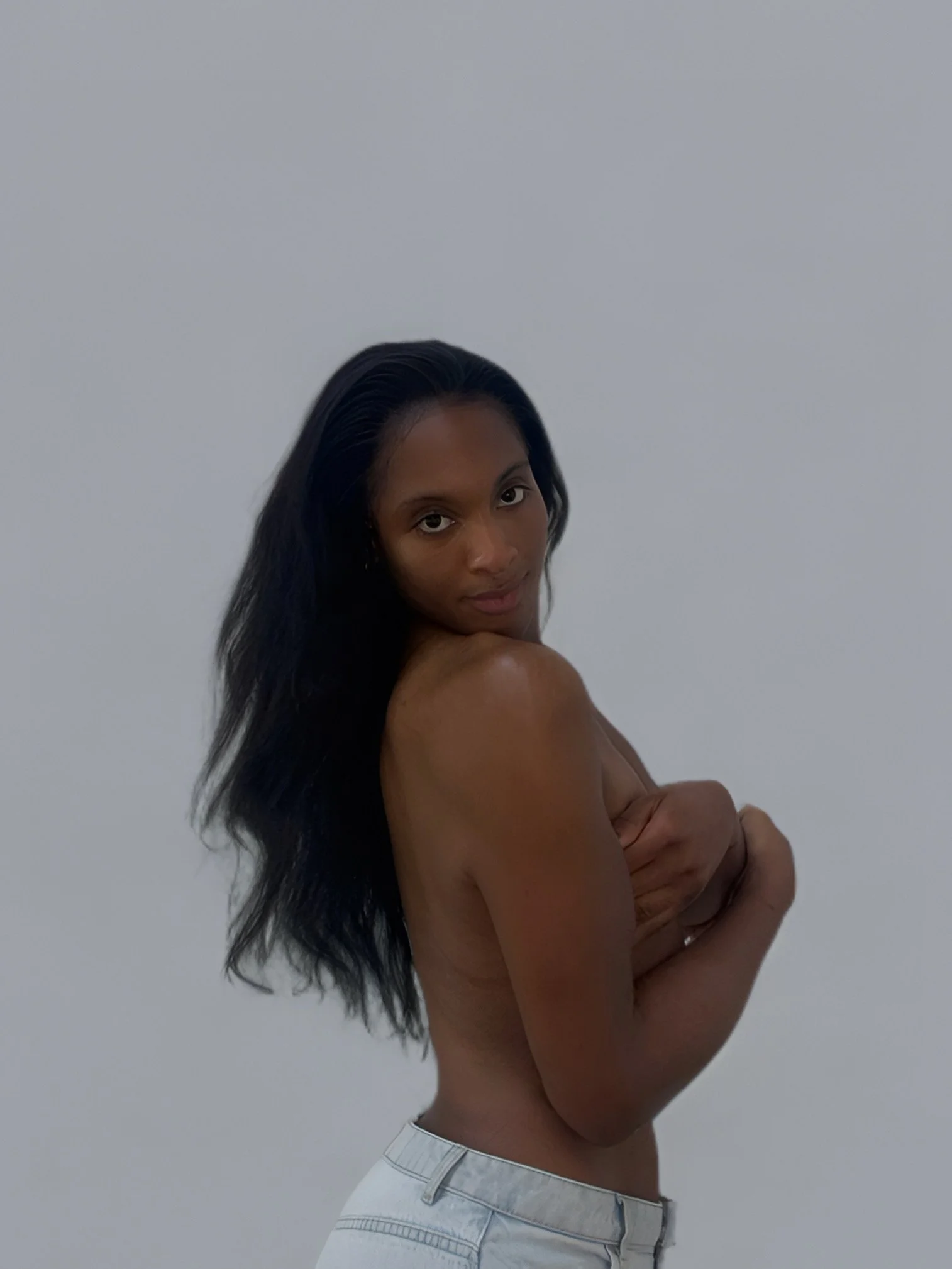 A woman with long black hair, topless, wearing white jeans, stands against a plain white wall with her arms crossed.