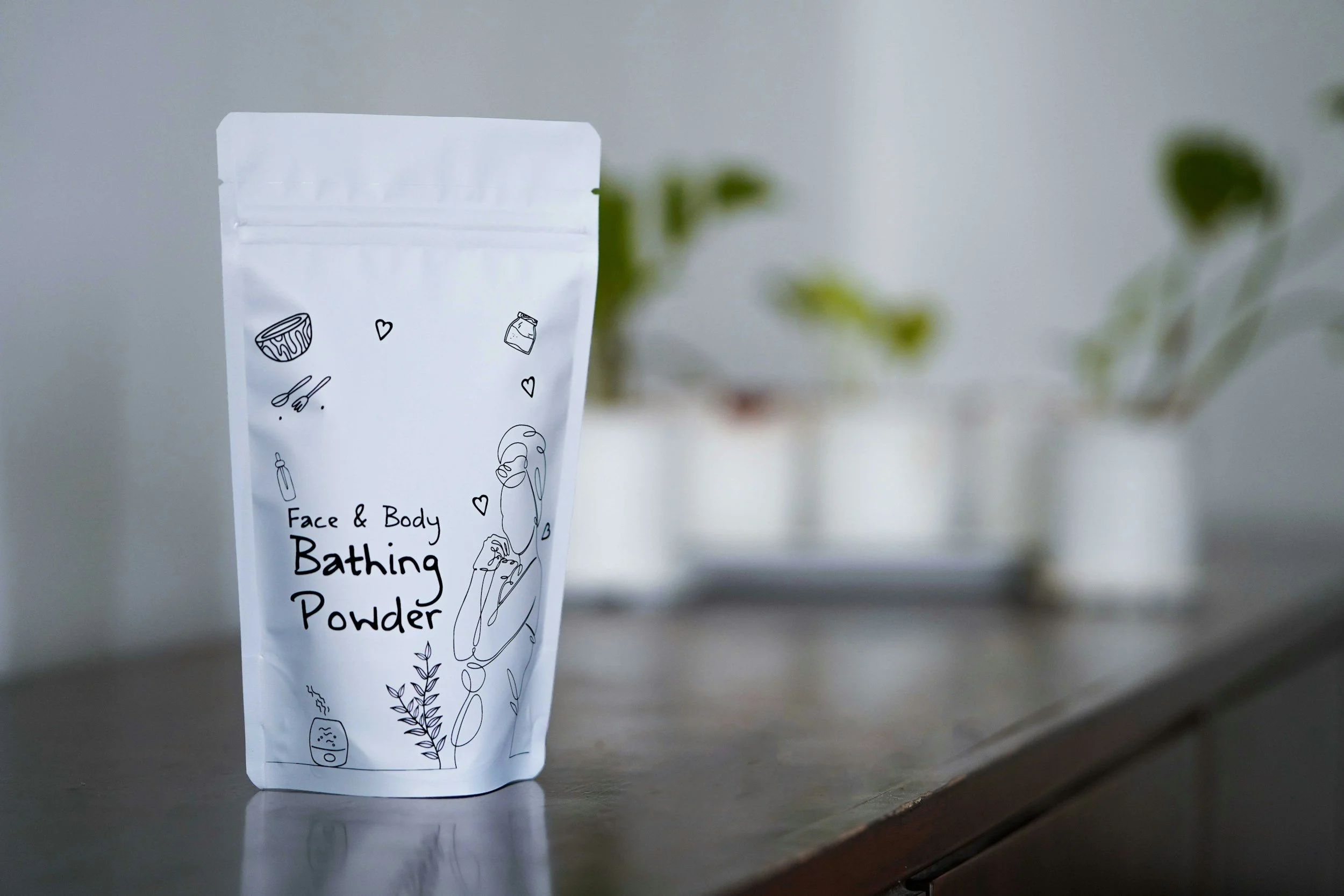 White stand-up pouch labeled "Face & Body Bathing Powder," decorated with black line drawings of a bowl, heart, spoon, jar, woman, and plants, placed on a metallic surface with a blurred green plant in the background.