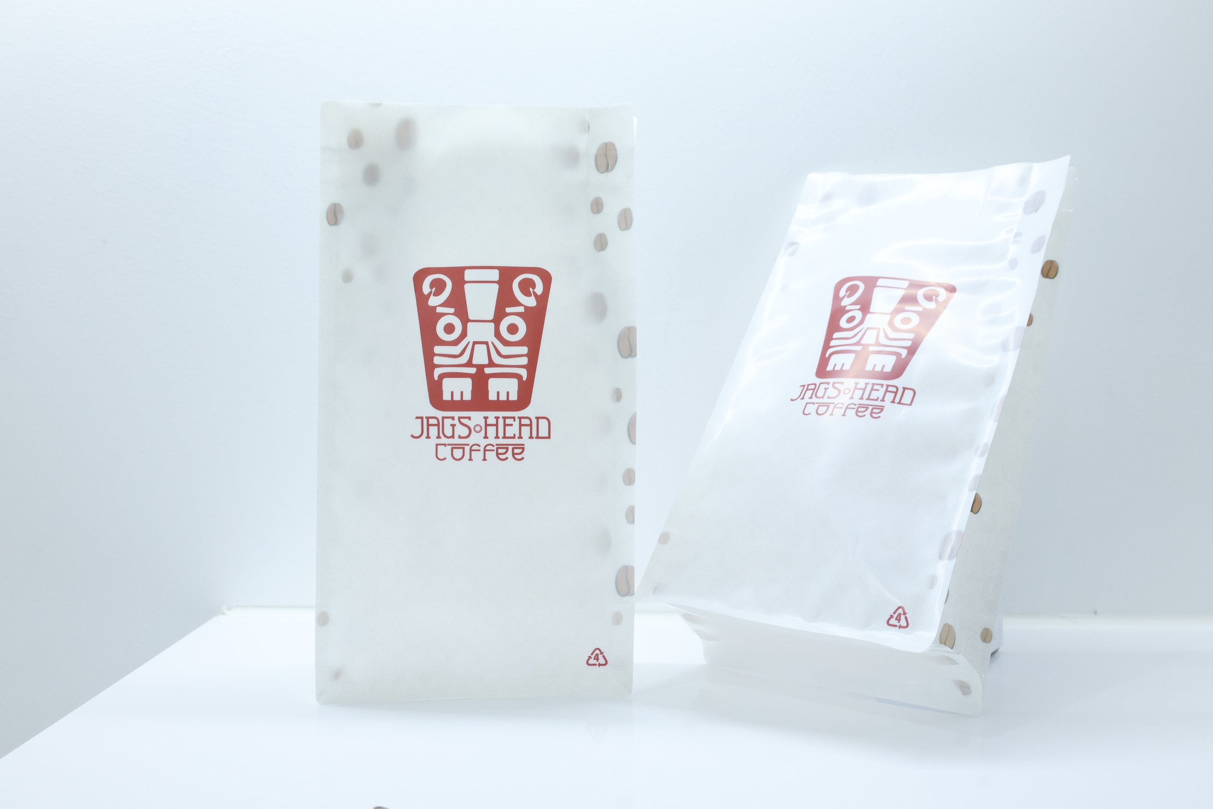 Two white paper bags with the Jag's Head Coffee logo, one in front of the other, and a white bag with a recycling symbol, all positioned on a white surface against a white background.