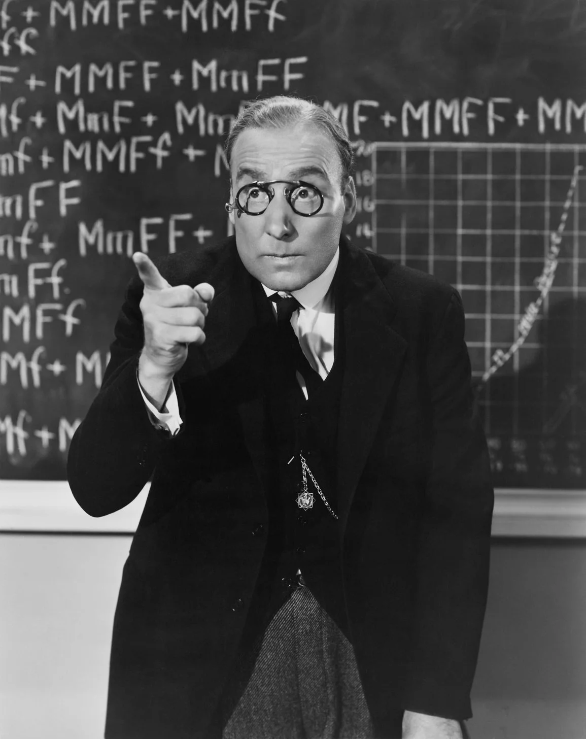 Homme en costume sombre et lunettes rondes devant un tableau noir couvert de formules, pointant du doigt comme pour expliquer une leçon.