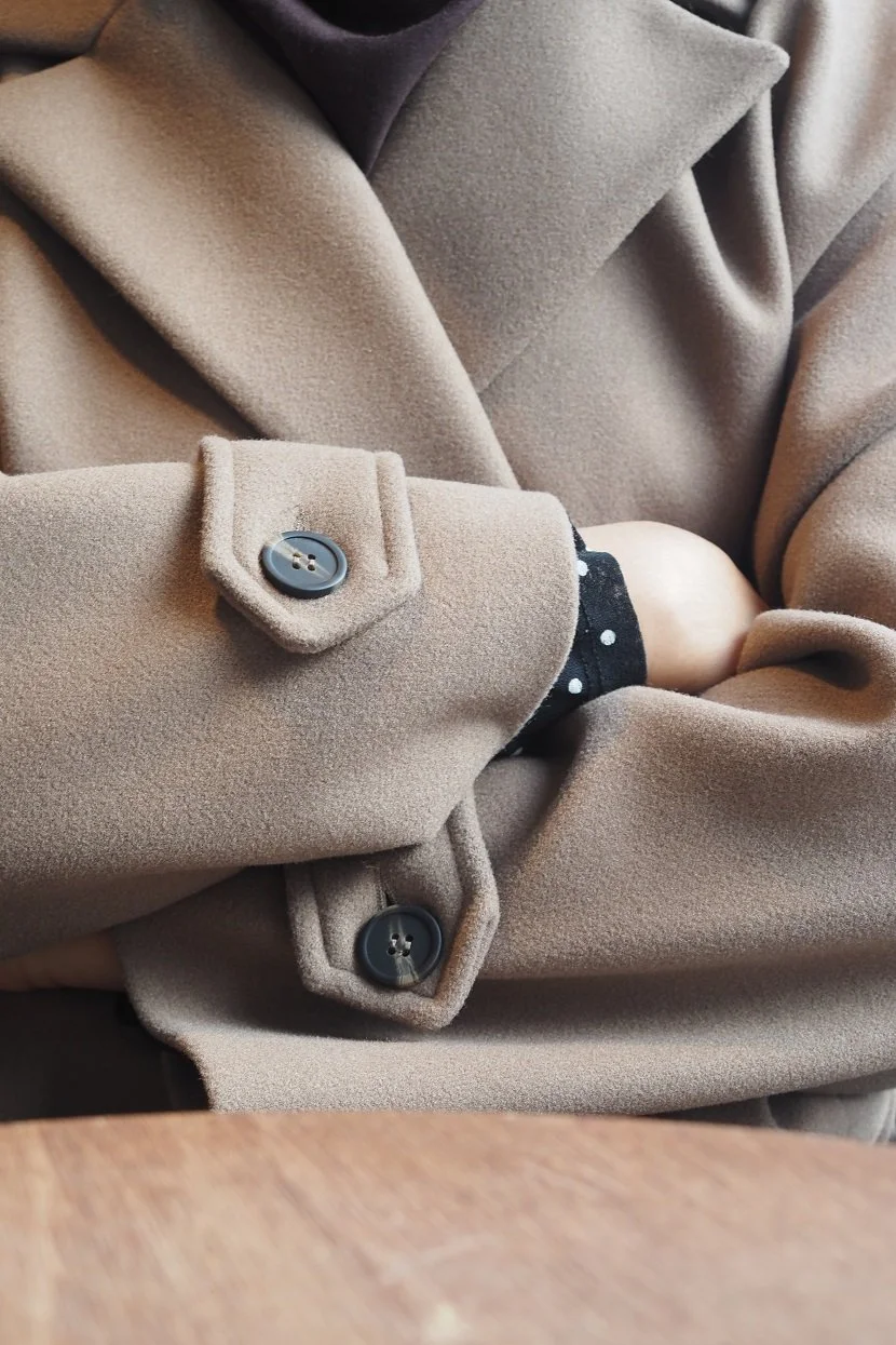 Gros plan sur un manteau beige en laine, bras croisés, montrant les boutons et les coutures du tissu. 