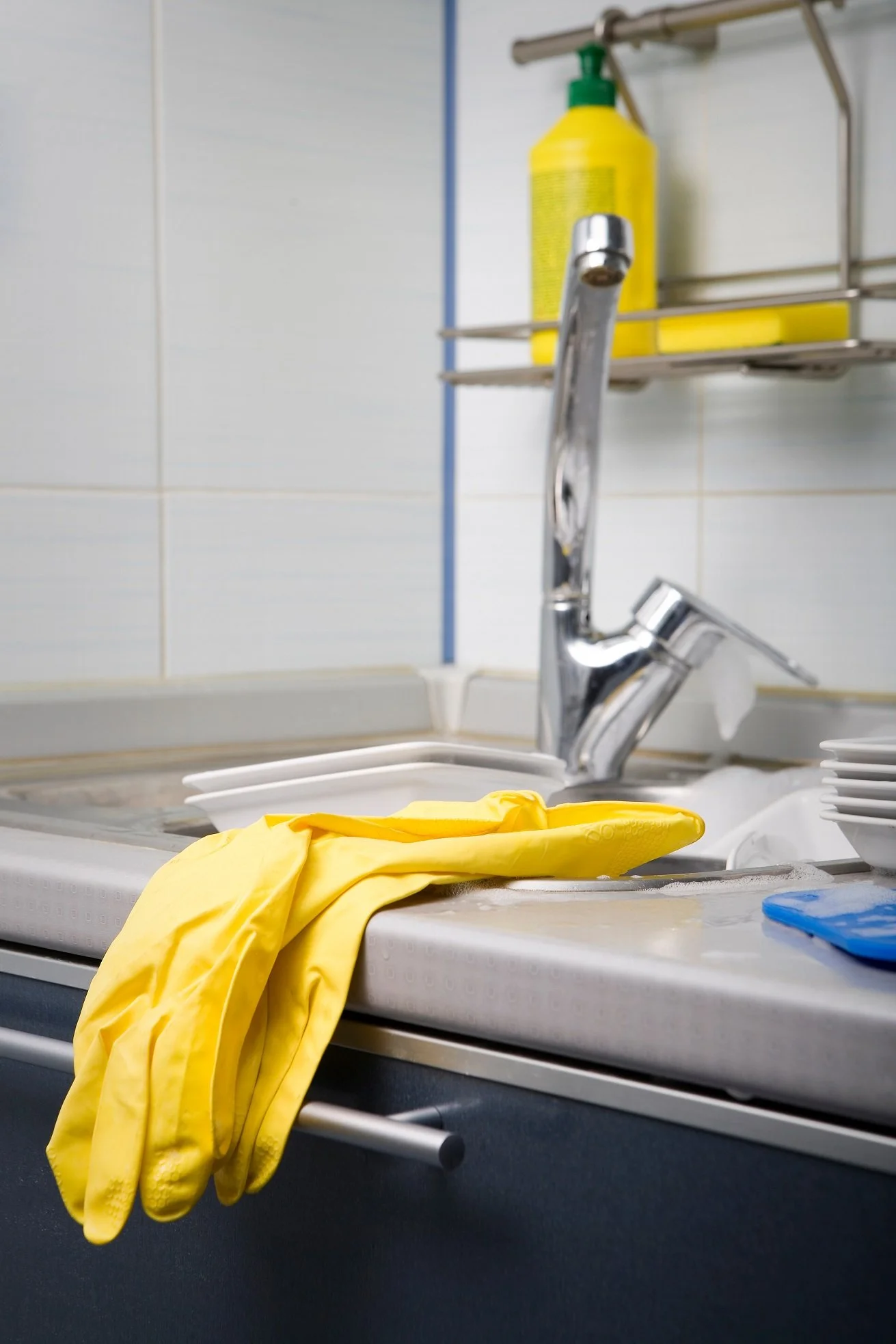 Évier de cuisine avec robinet chromé, paire de gants de nettoyage jaunes posée sur le bord, bouteille de liquide vaisselle et vaisselle blanche empilée à côté.