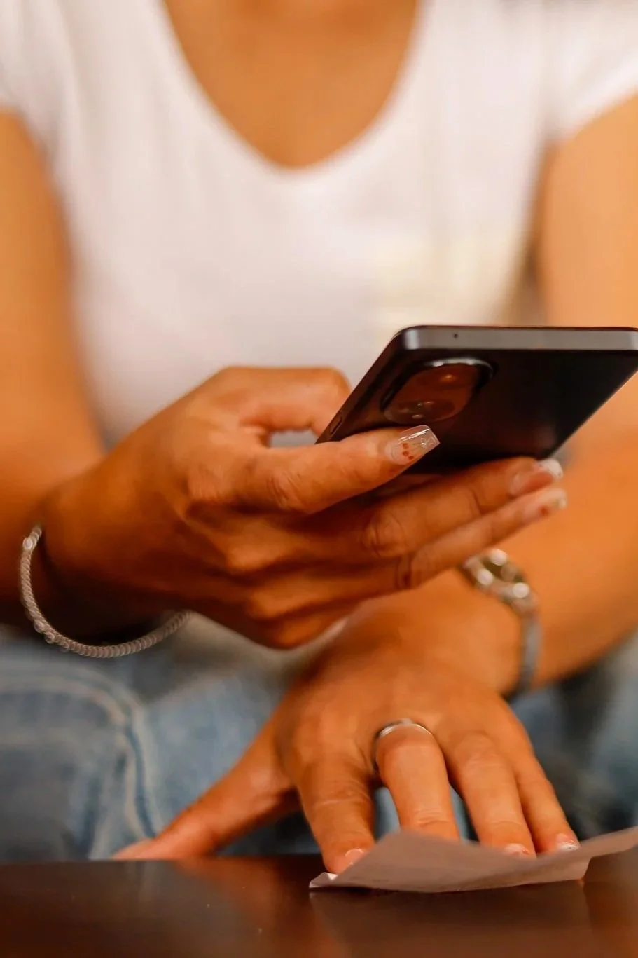 Main textant sur un téléphone portable, l’autre posée sur une note en papier ; personne vêtue d’un haut blanc et d’un jean.