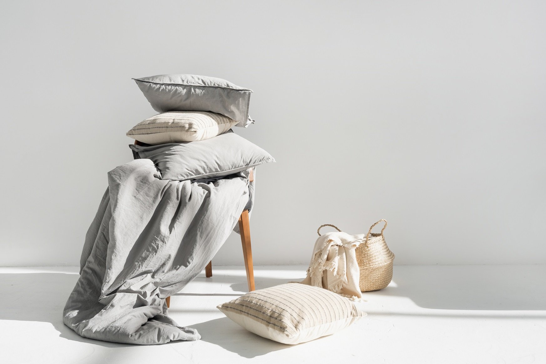 Empilement de coussins et de linge de maison gris clair sur un tabouret en bois, avec deux sacs tressés posés au sol dans une lumière blanche et apaisante.