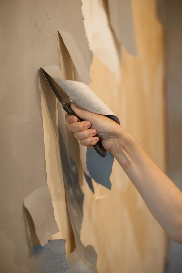 Main tenant un grattoir en métal, retirant du papier peint beige d’un mur pour révéler les couches sous jacentes.