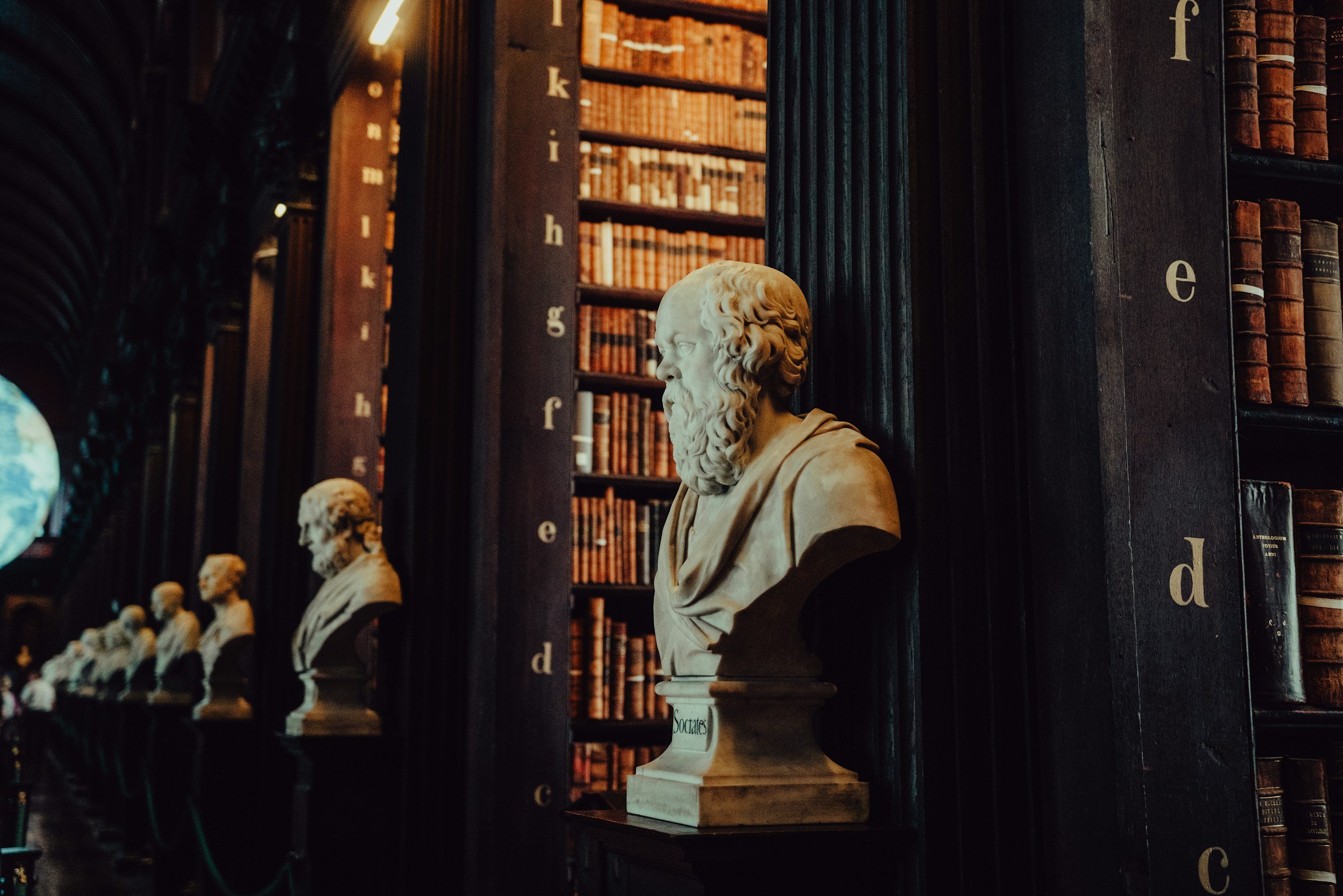 Sculpted busts of ancient philosophers lined up along stacked books in a dimly lit library.