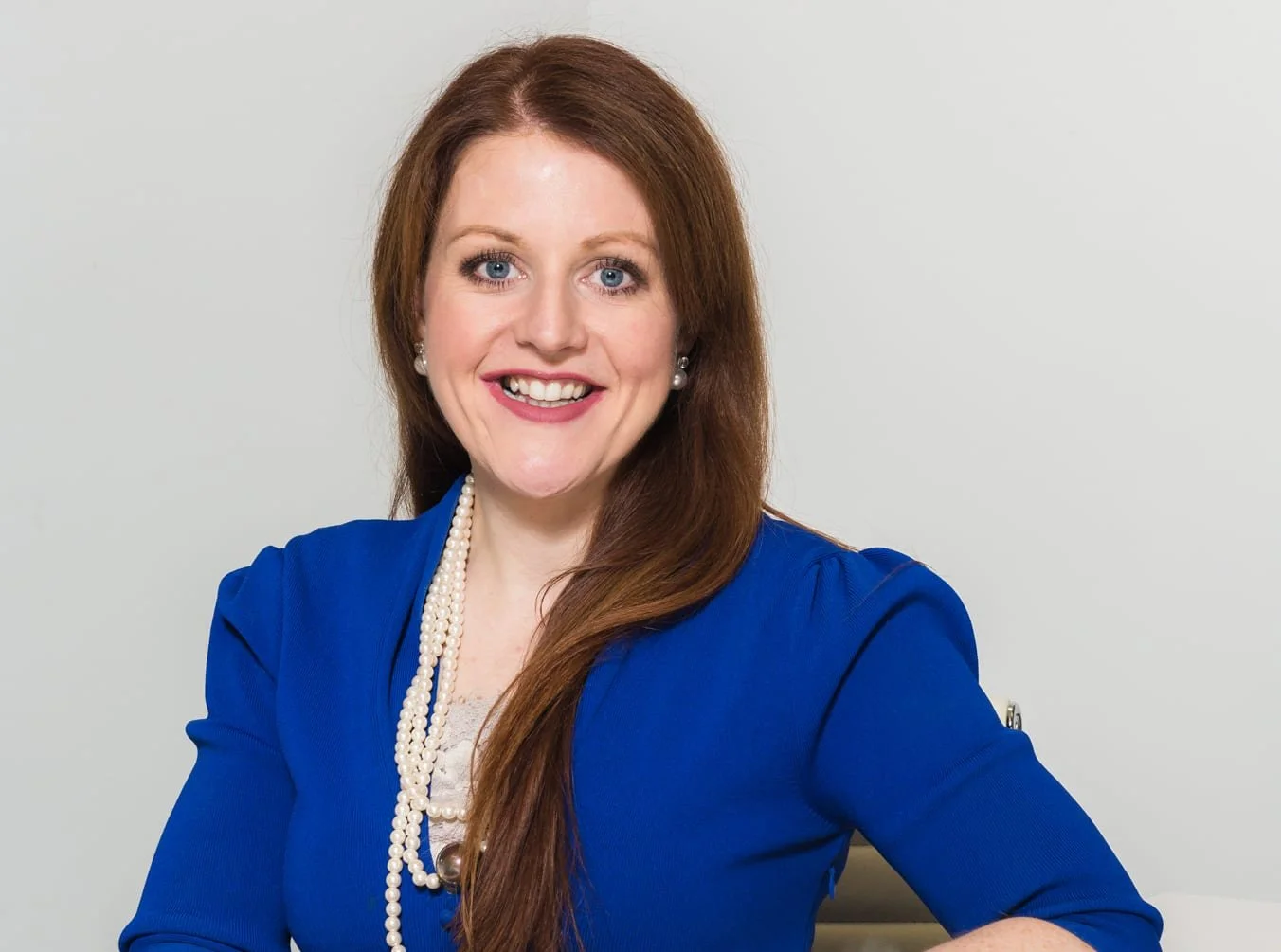 A woman with long red hair wearing a blue blazer, pearl earrings, and a pearl necklace, smiling against a plain light gray background.
