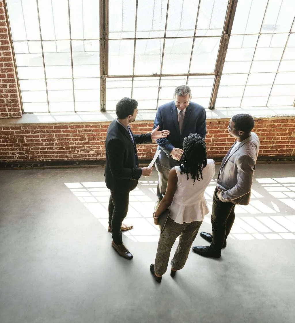 A group of four diverse professionals having a discussion in a spacious, industrial-style office with large windows and exposed brick walls.
