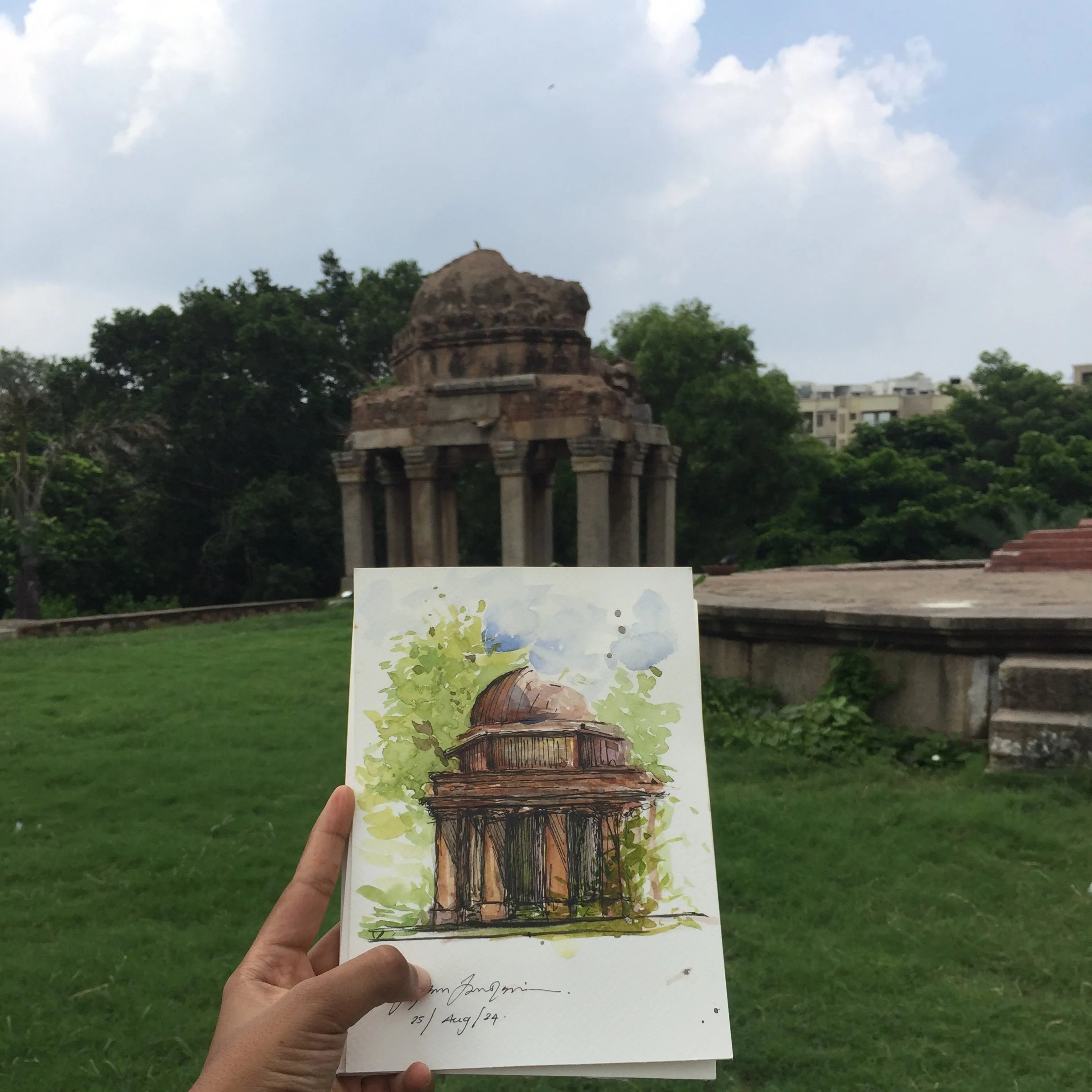 A person holding a watercolor painting of a temple with greenery, in an outdoor park setting with trees and buildings in the background.