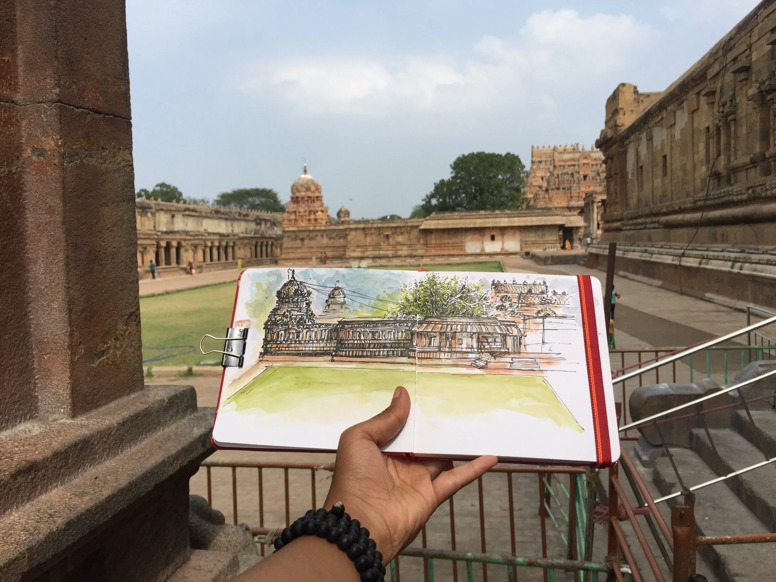 A person holding an open sketchbook with a drawing of an old temple complex, set against a historic stone building and temple ruins in the background.