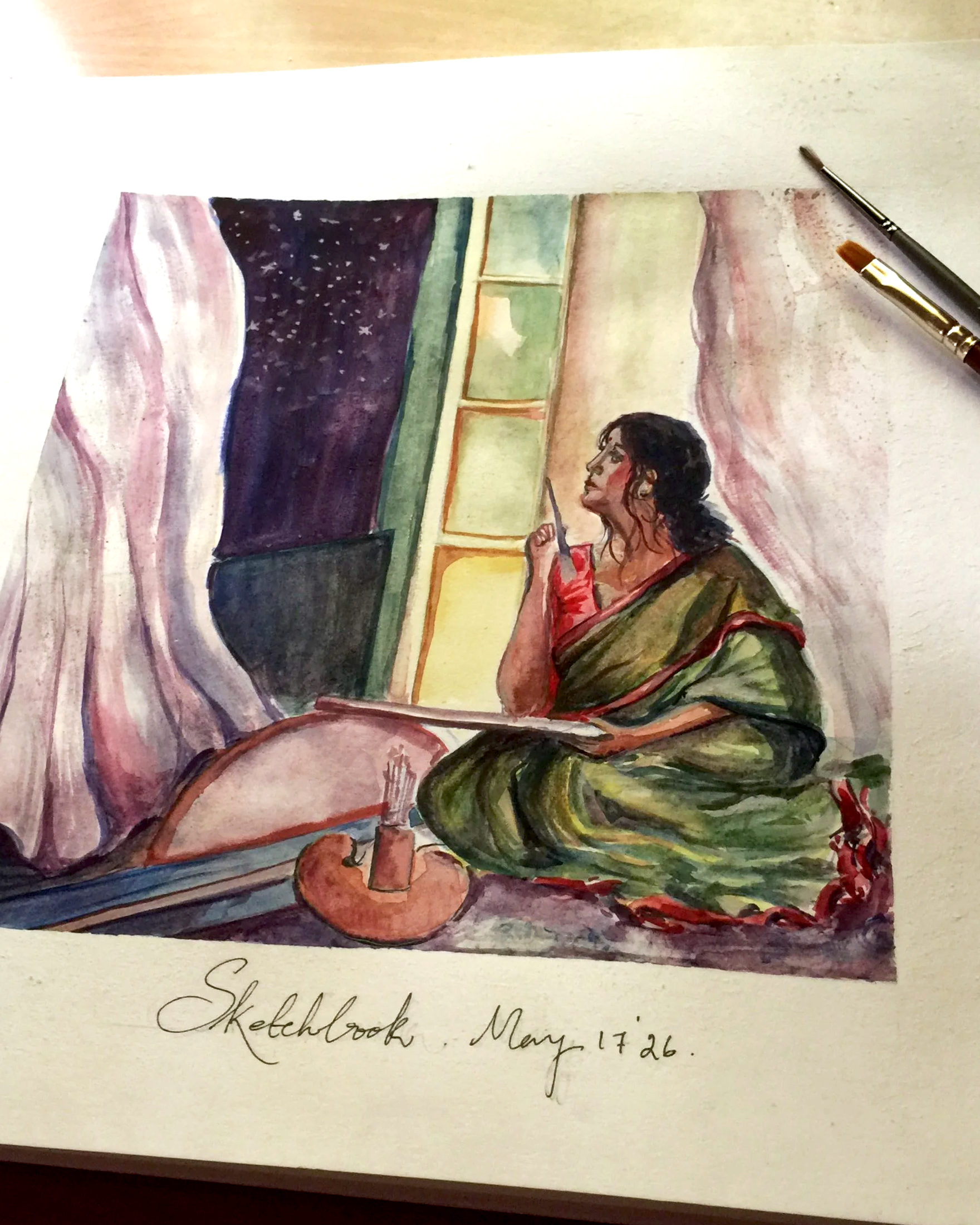 Watercolor painting of a woman with dark hair wearing a green and red dress, sitting on the floor, in front of a window with a night sky full of stars; the painting is labeled "Sketchbook, May 17 26".