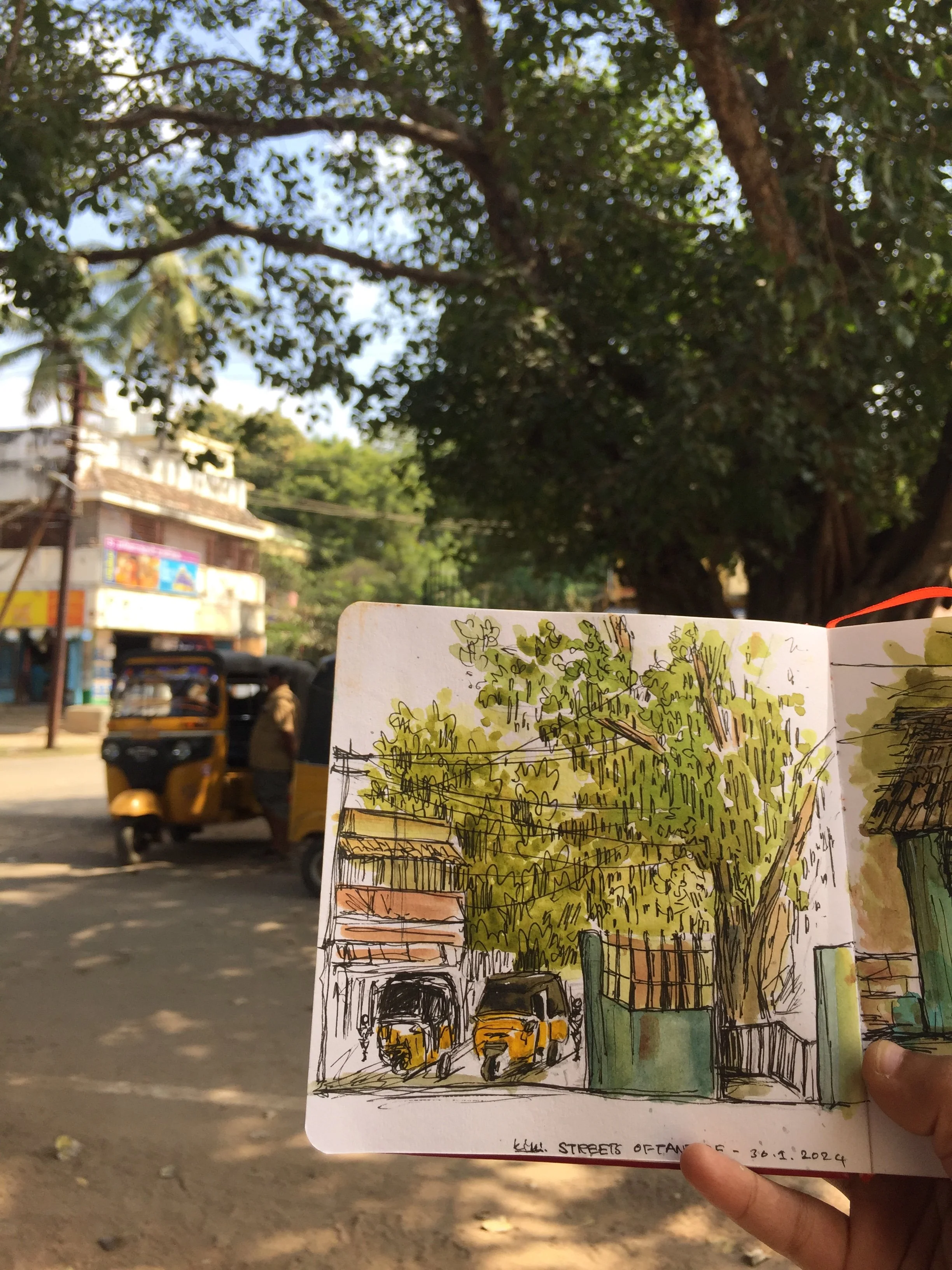 A hand holding a sketchbook with a colorful drawing, partially covering a busy street scene with auto rickshaws and a large tree in the background.