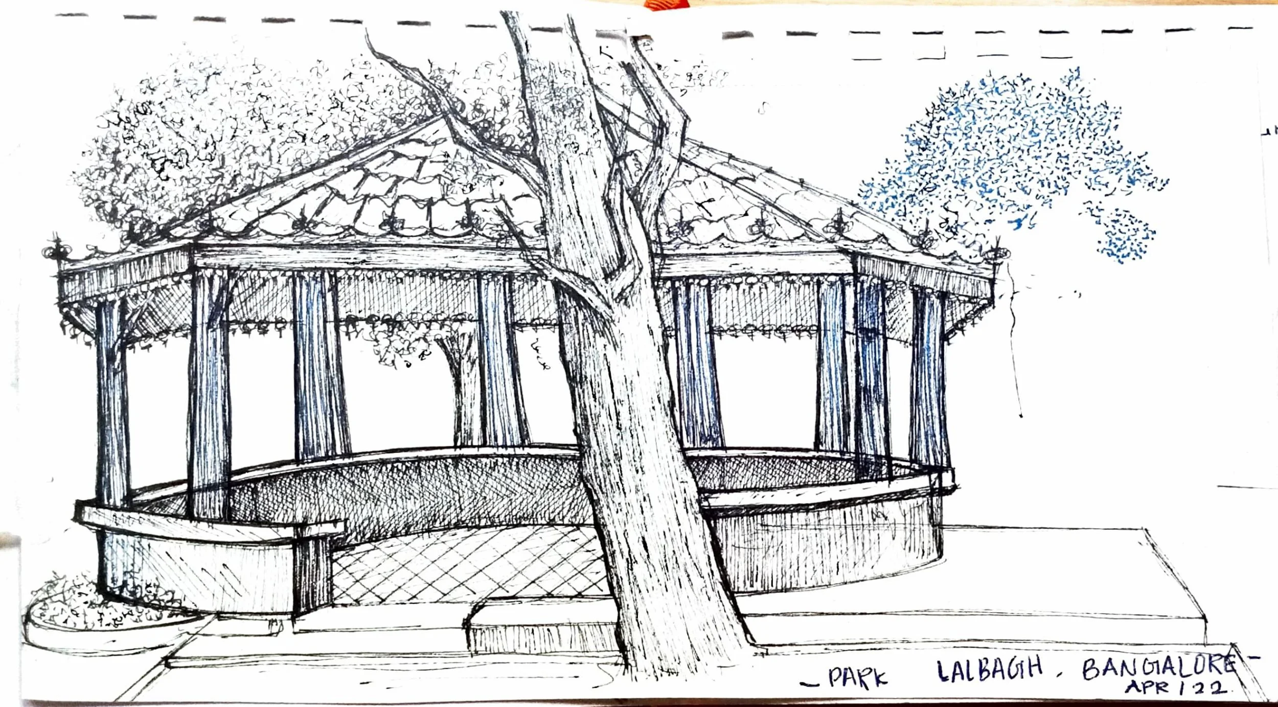 A detailed sketch of a pavilion or gazebo structure in a park, with a large tree growing through the middle. The gazebo has a tiled roof, decorative posts, and string lights hanging inside. There are small plants and trees around the structure, with 
