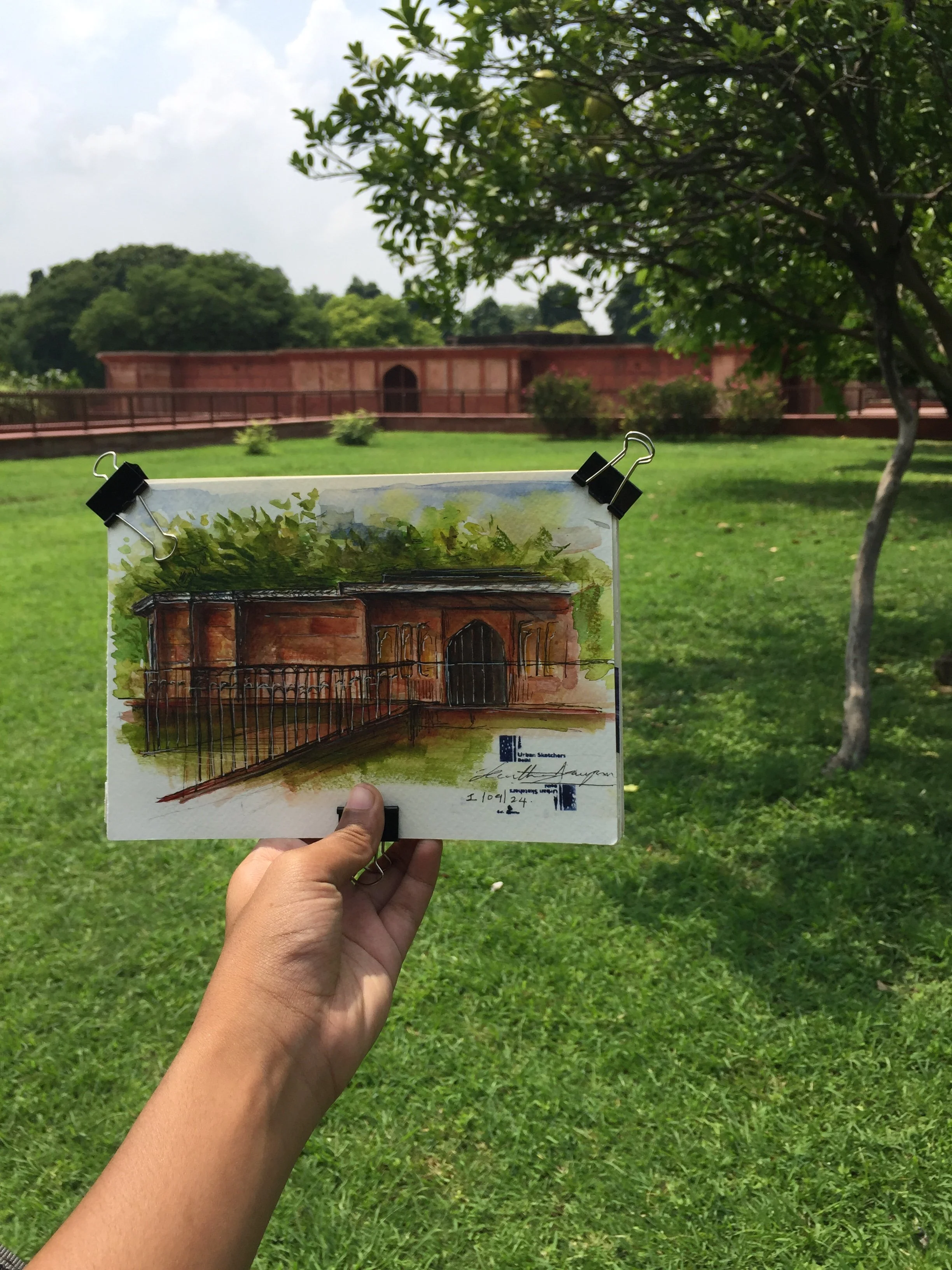Hand holding a postcard painted with an architectural sketch of a brick building with arched doorways, set in a green outdoor environment.