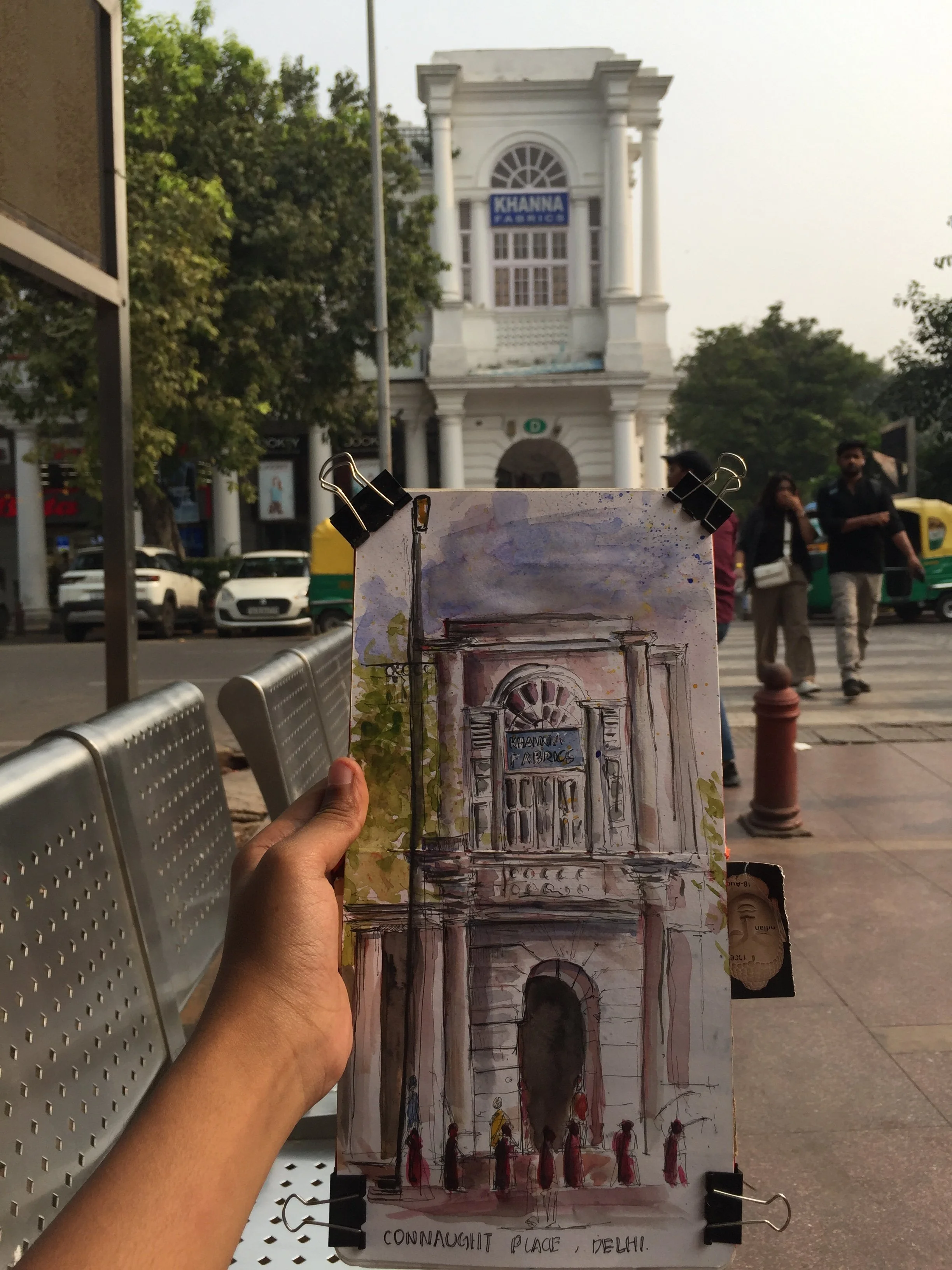 A hand holding a watercolor painting of Connaught Place in Delhi, India, with a historic white building in the background featuring large pillars and an arched window, and people walking nearby.